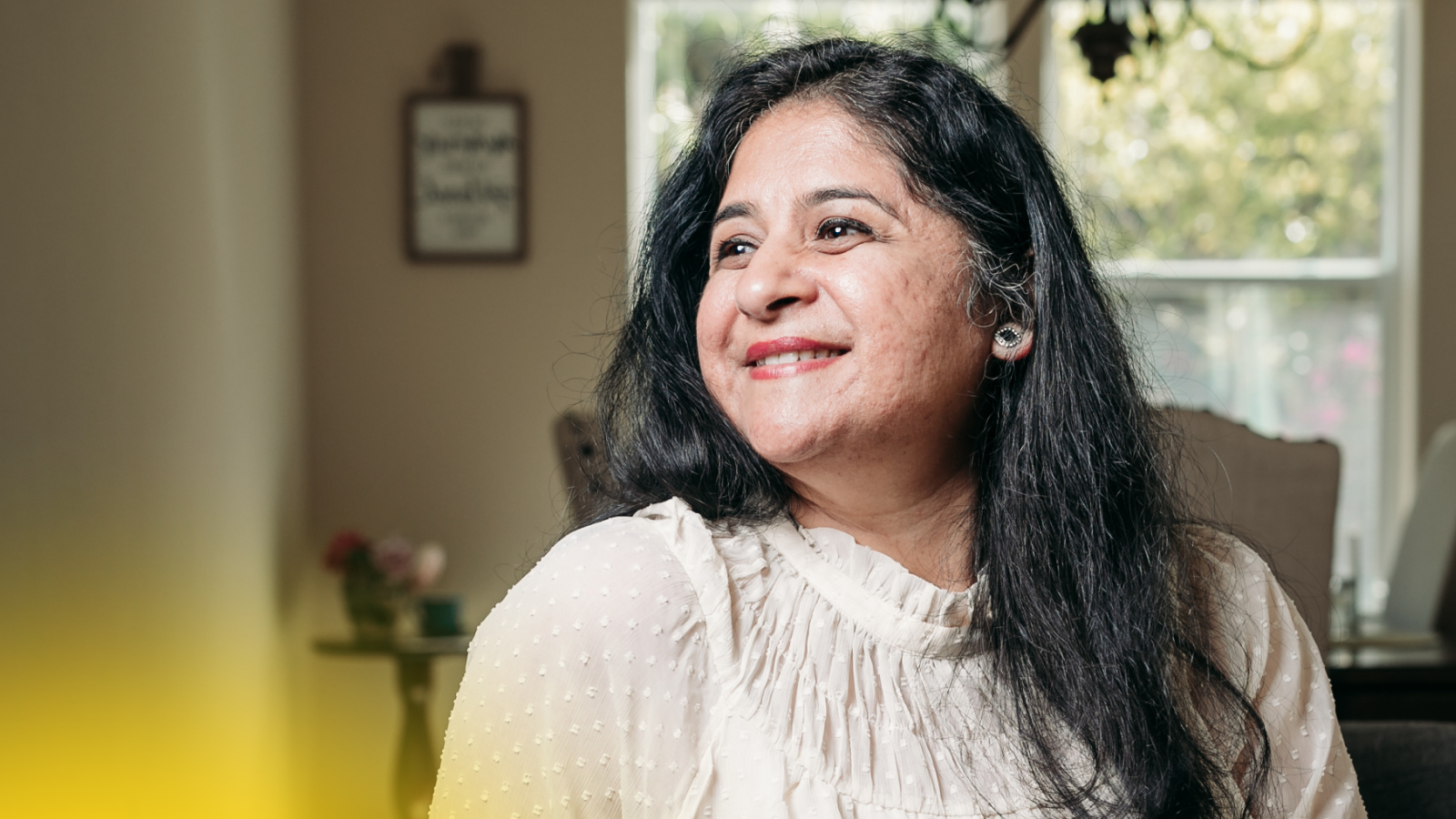 A middle-aged woman with long, dark hair looks to the side and smiles, bathed in natural light from a nearby window.