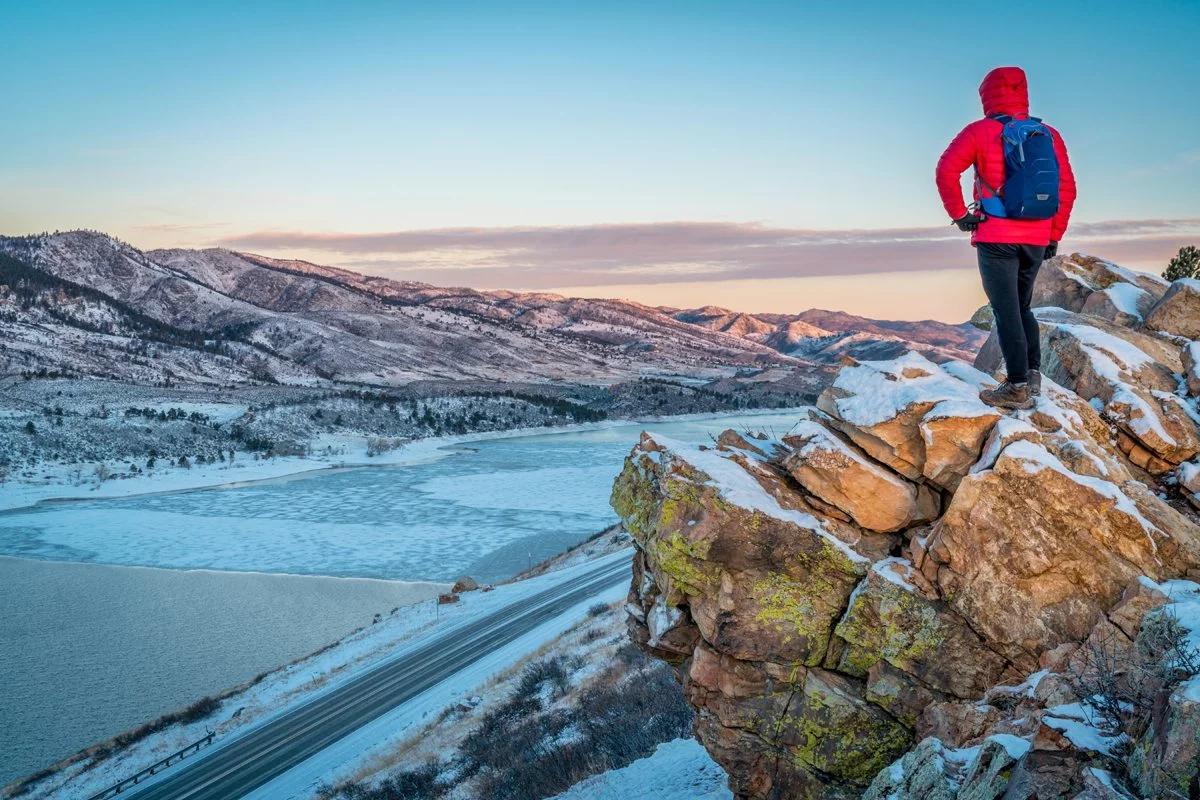 Person wearing hiking boots and a backpack standing on top of a tall rock cliff looking out over mountains