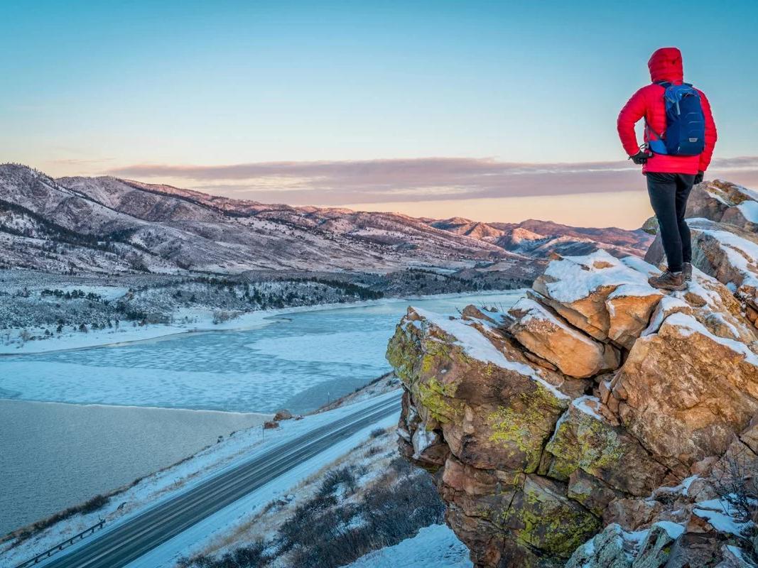 Person wearing hiking boots and a backpack standing on top of a tall rock cliff looking out over mountains