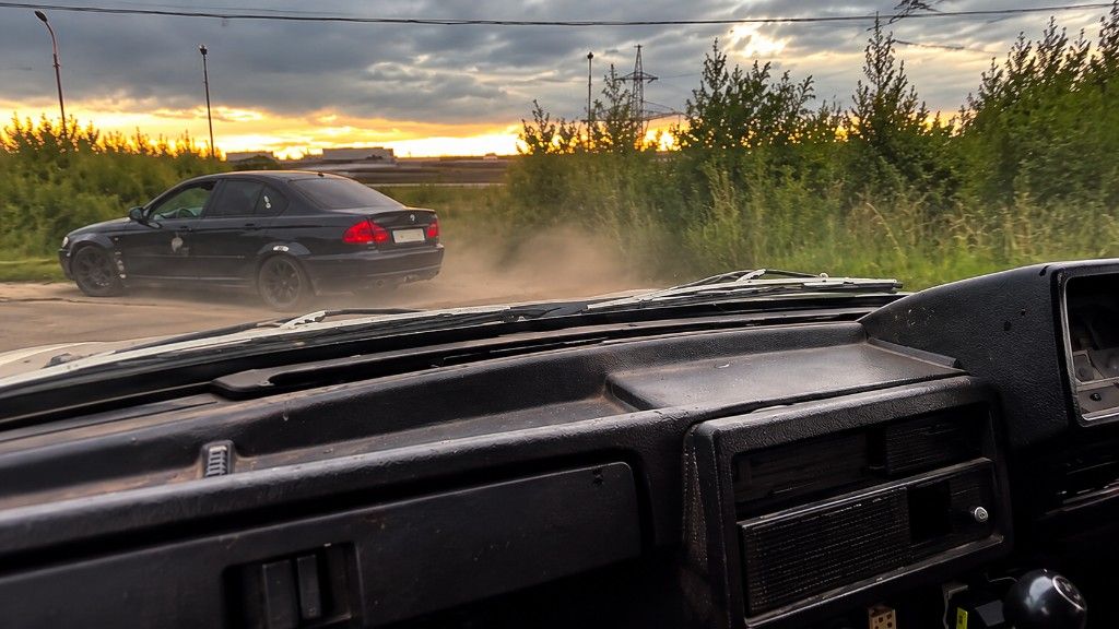 Pogled iz kabine: crni automobil diže prašinu na sporednom putu, zalazak sunca iza grmlja i dalekovoda.
