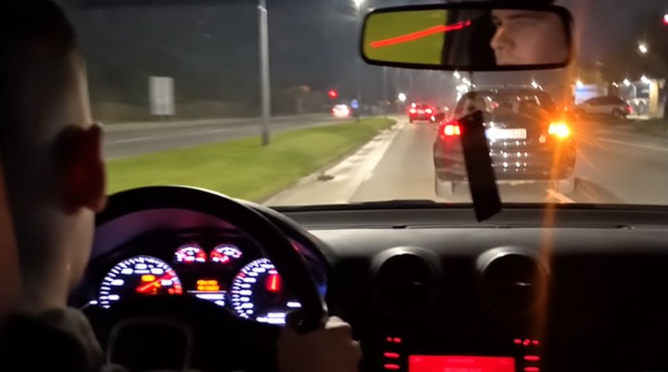 Noćna vožnja iz kabine: vozač s leđa, volan, instrument tabla i auto ispred. Lice se vidi u retrovizoru.