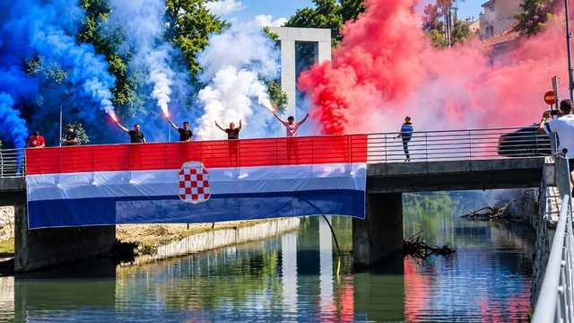Most preko rijeke, preko ograde visi crveno-bijelo-plava zastava sa šahovnicom. Na mostu ljudi drže baklje, dim je plavi, bijeli i crveni.