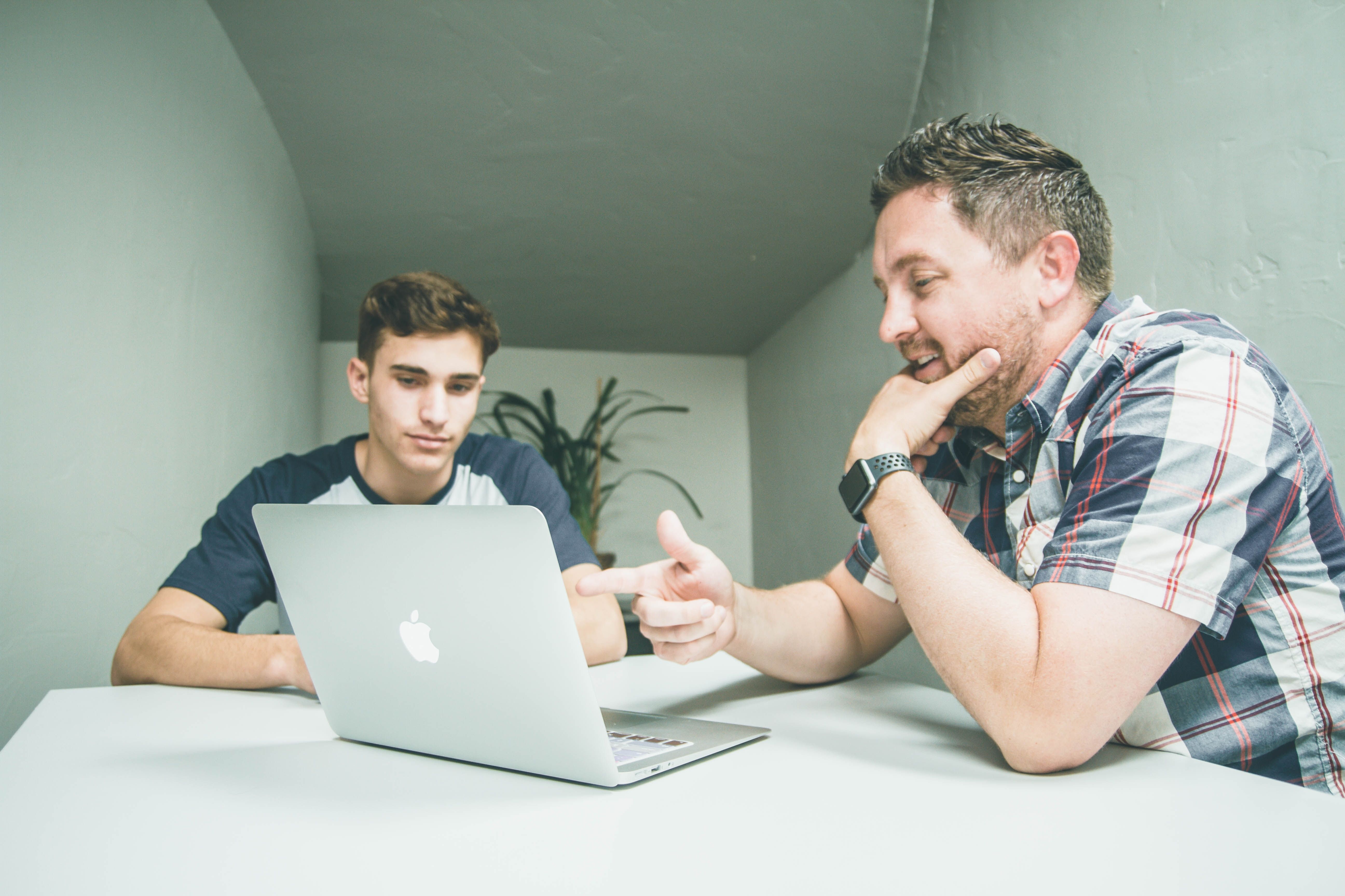 man wearing white and black plaid button-up sports shirt pointing at a silver MacBook