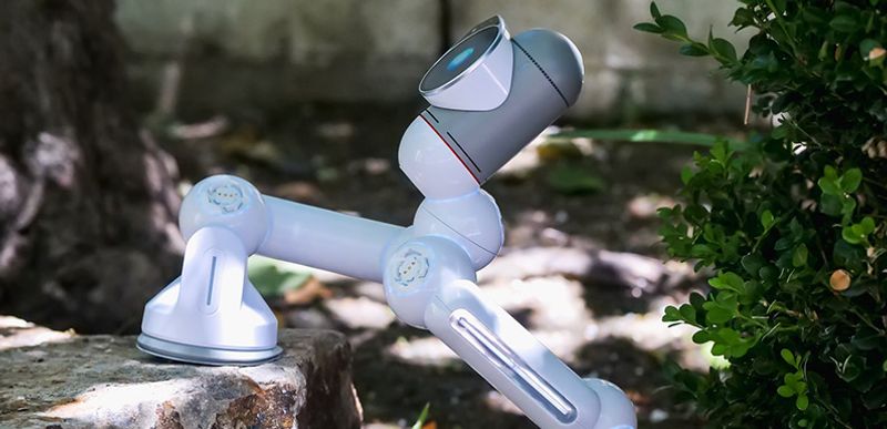 Photo of a white robotic arm with articulated joints and a camera-like sensor head, mounted on a rock outdoors.