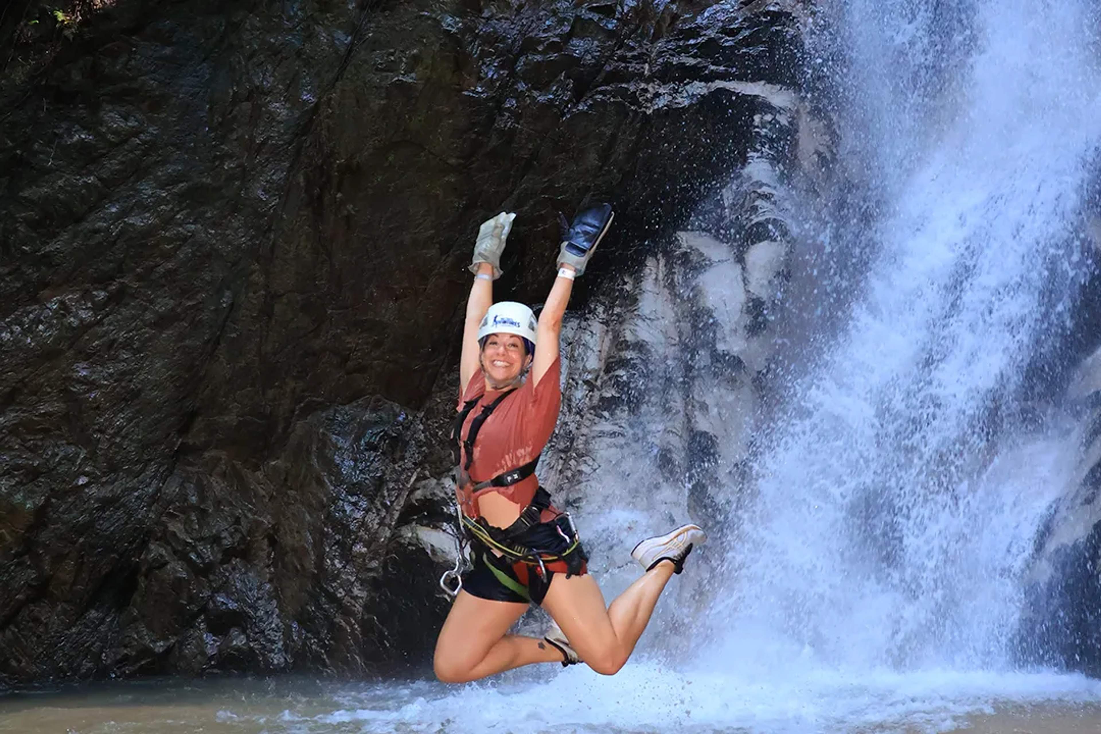 Mujer salta con alegría frente a una cascada en una aventura al aire libre.