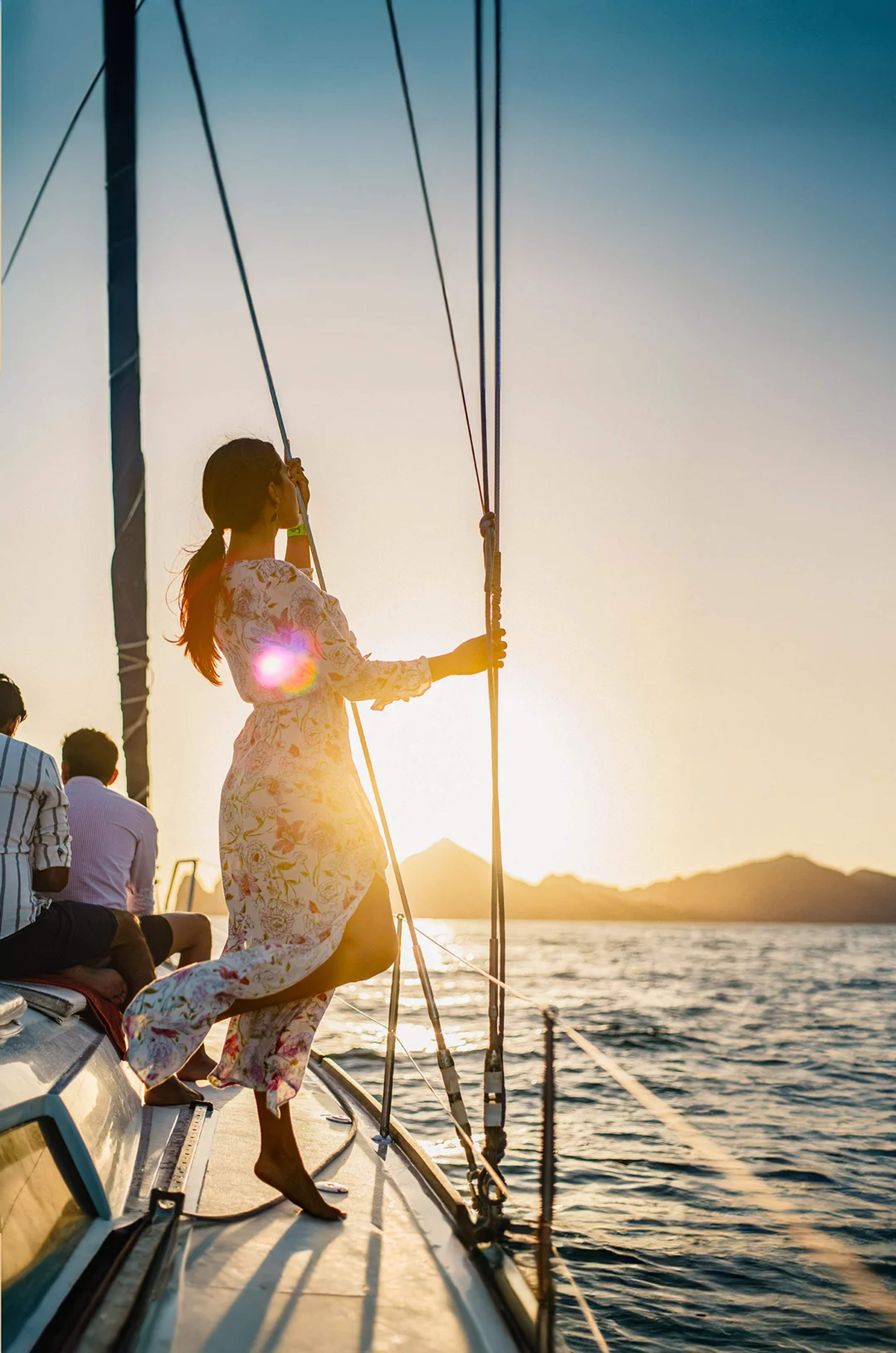 Mujer disfruta del atardecer en un velero, mirando el mar con montañas al fondo.