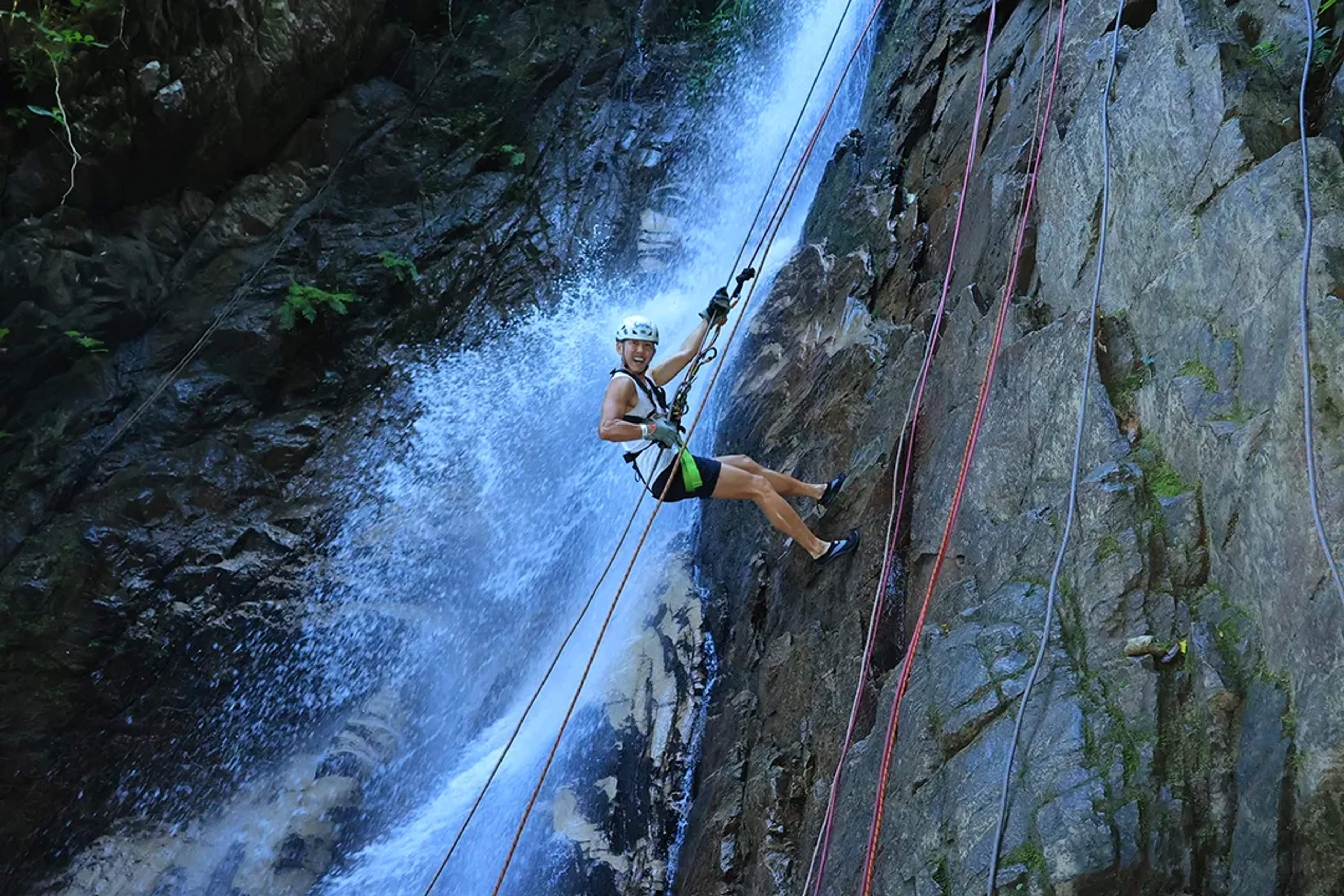 Mujer haciendo rappel en una cascada, sonriendo entre la bruma y acantilados rocosos.