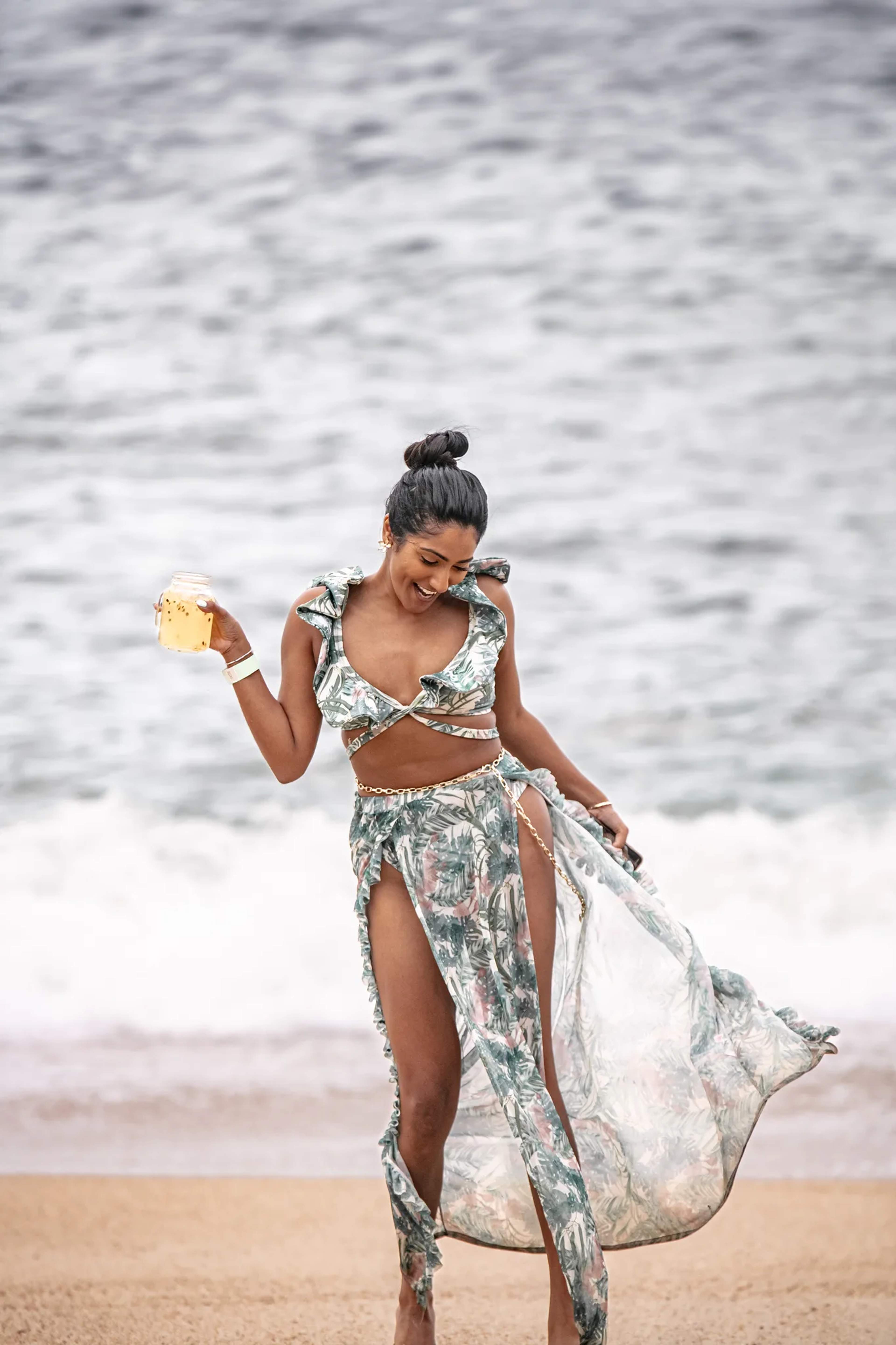A woman on a tropical beach holds a refreshing drink, wearing a patterned two-piece outfit and a sheer cover-up.