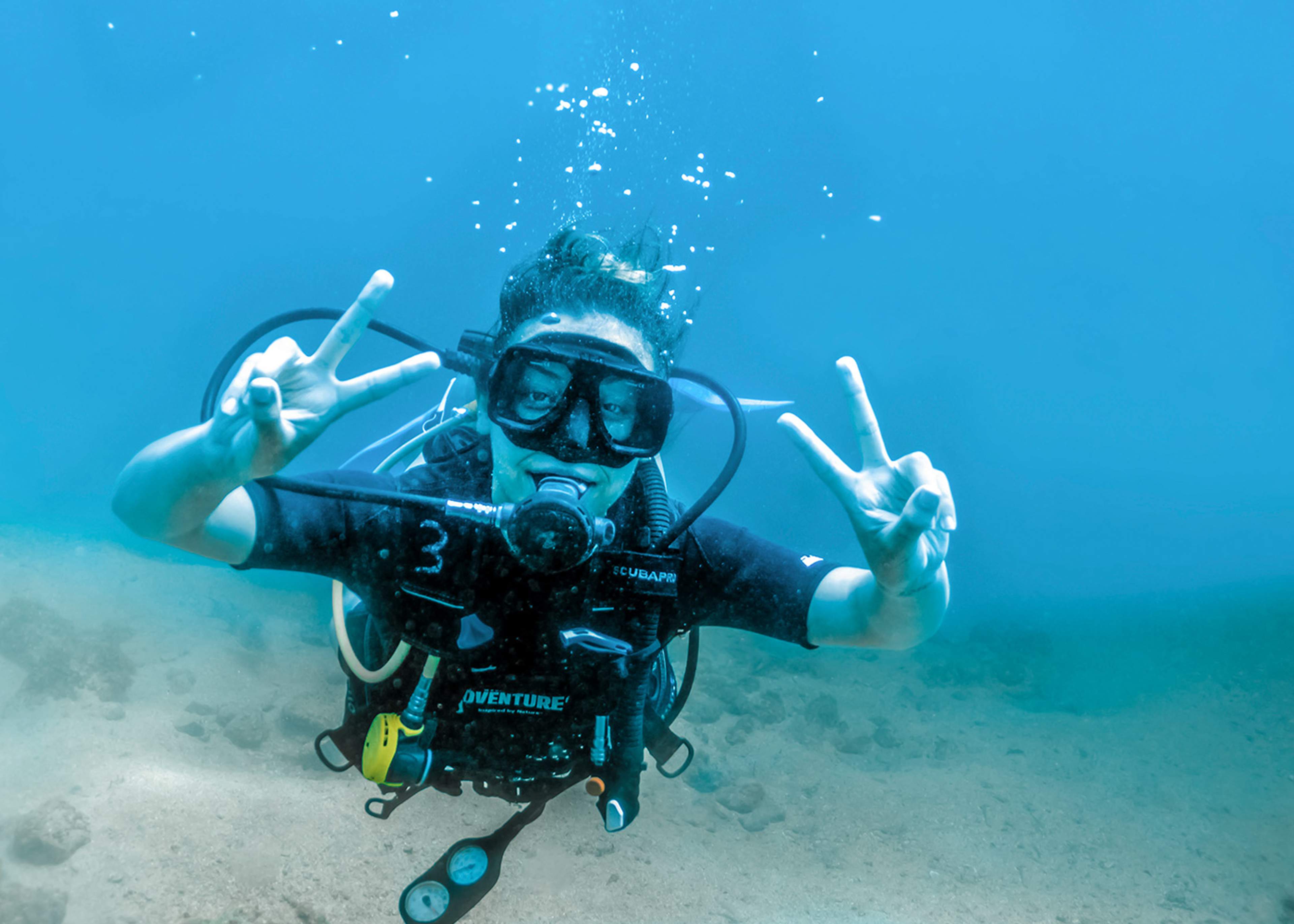 A scuba diver underwater making peace signs with both hands, enjoying a dive in clear blue waters.