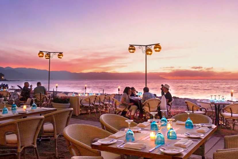 Oceanfront sunset dining experience with candlelit tables, seaside restaurant ambiance and guests enjoying dinner by the sea.