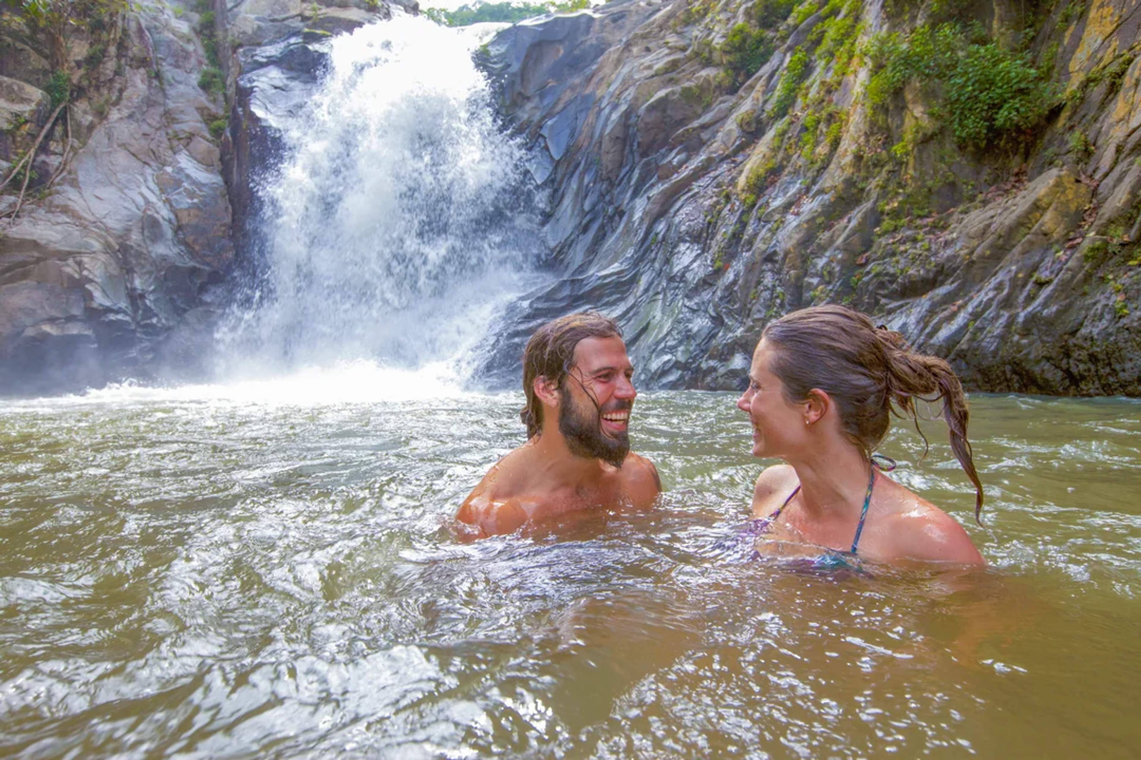 A couple enjoys a refreshing swim at the base of a jungle waterfall, smiling and surrounded by nature.