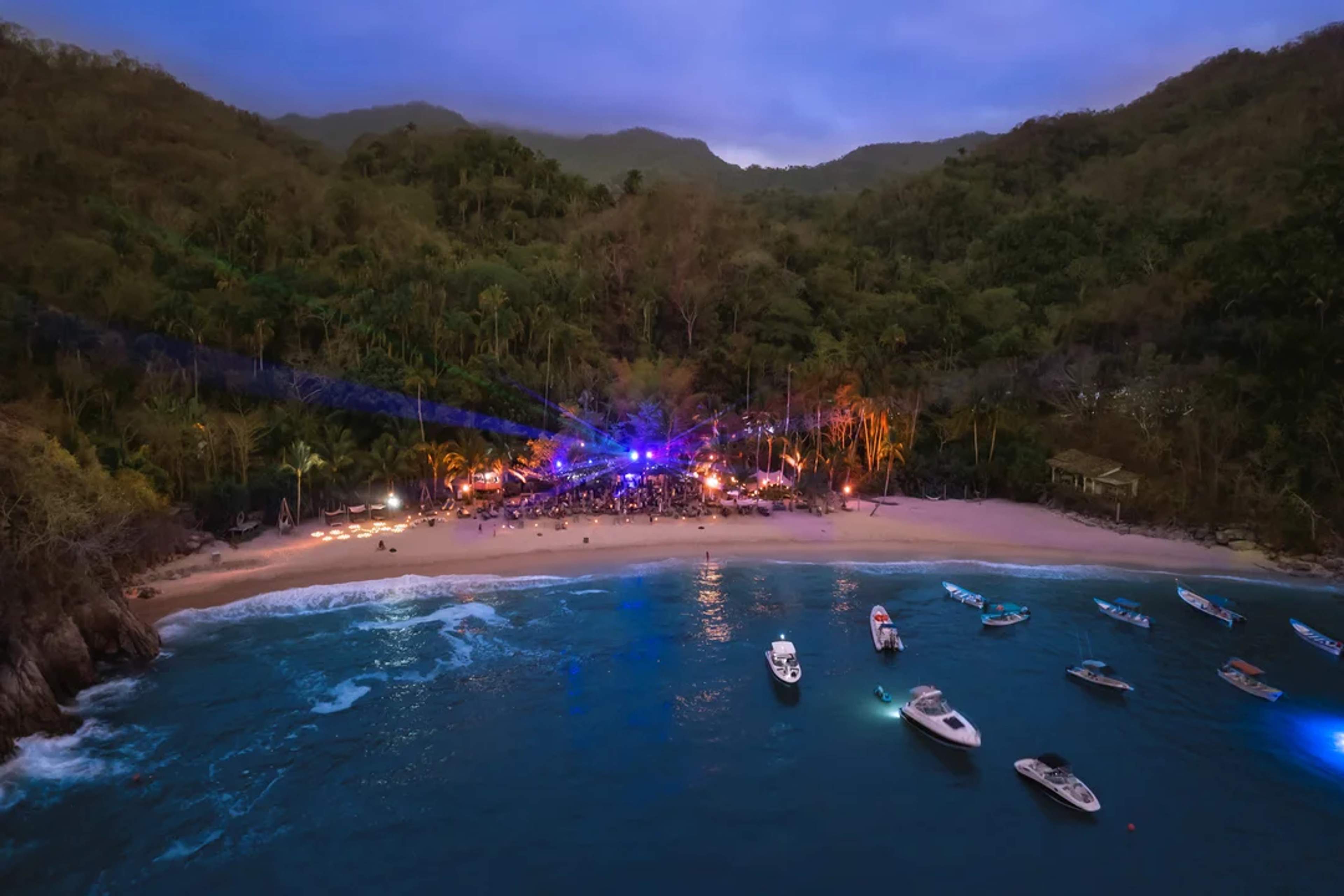 Fiesta nocturna en playa escondida, rodeada de selva, con luces y botes anclados en el mar.