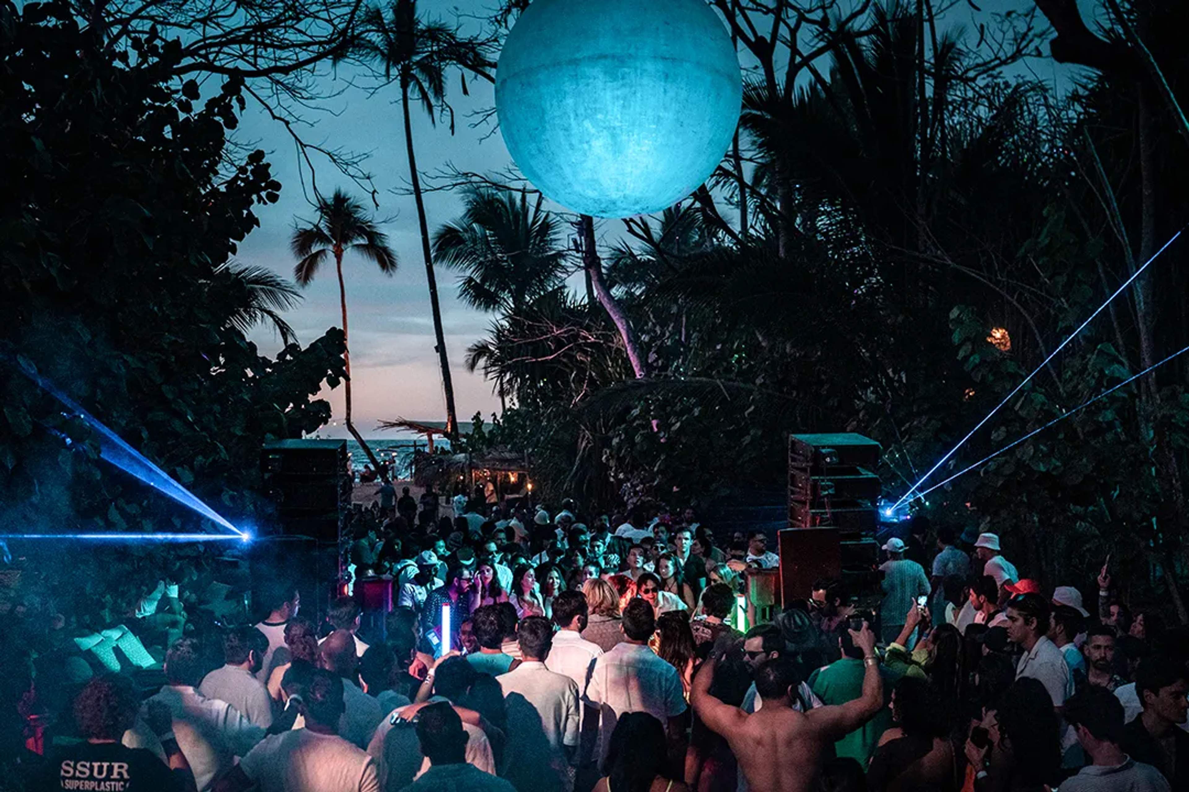 Beach party at night with lights, music, palm trees, and a crowd dancing under a glowing orb.