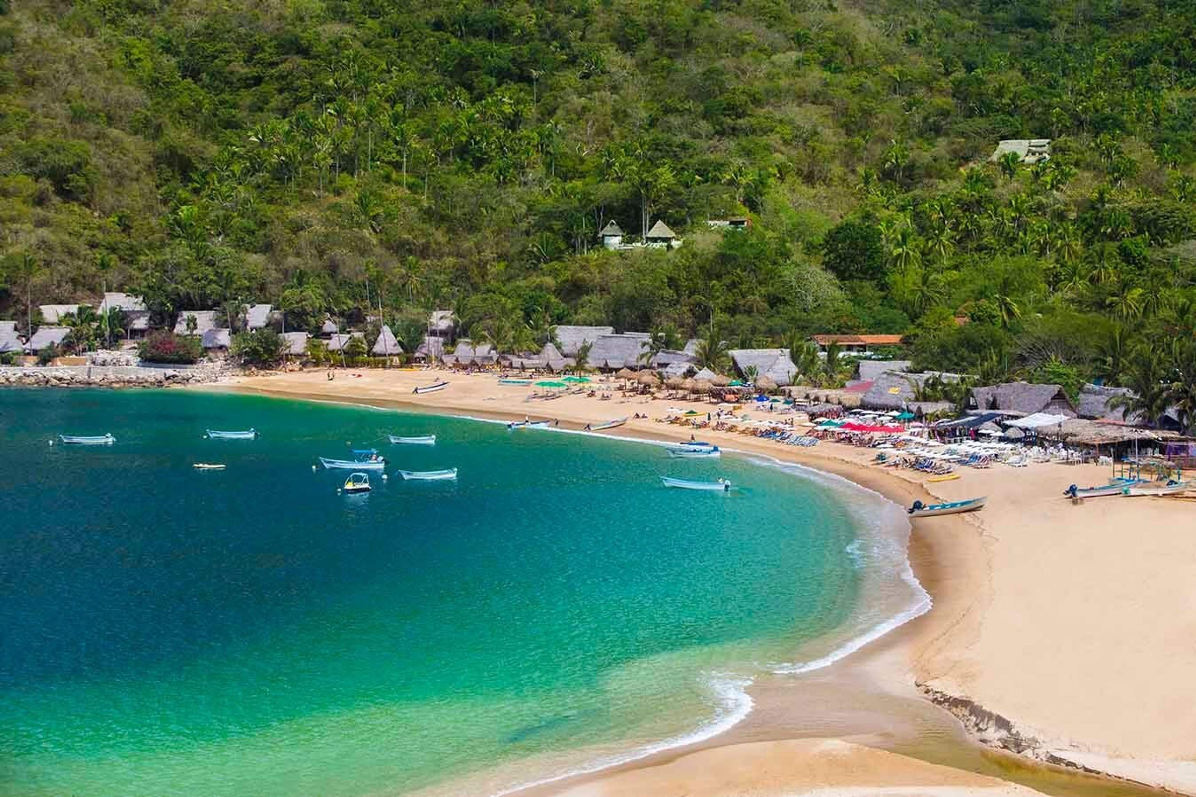 Playa tropical con aguas turquesas, barcos anclados, cabañas con techo de paja, sombrillas de playa y vegetación exuberante al fondo.