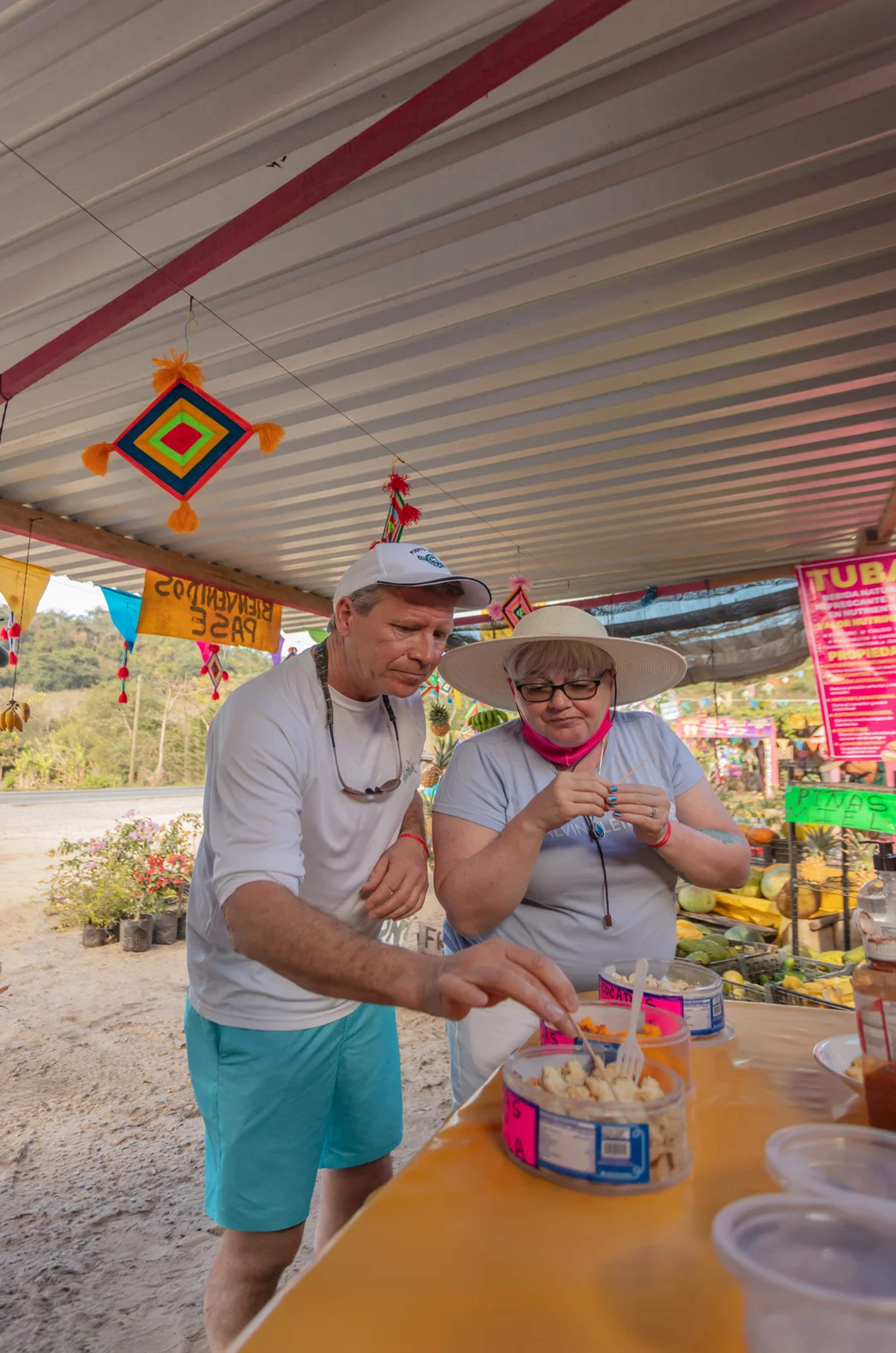 Tourists sampling local snacks at a colorful roadside market filled with fruits and handmade decor.