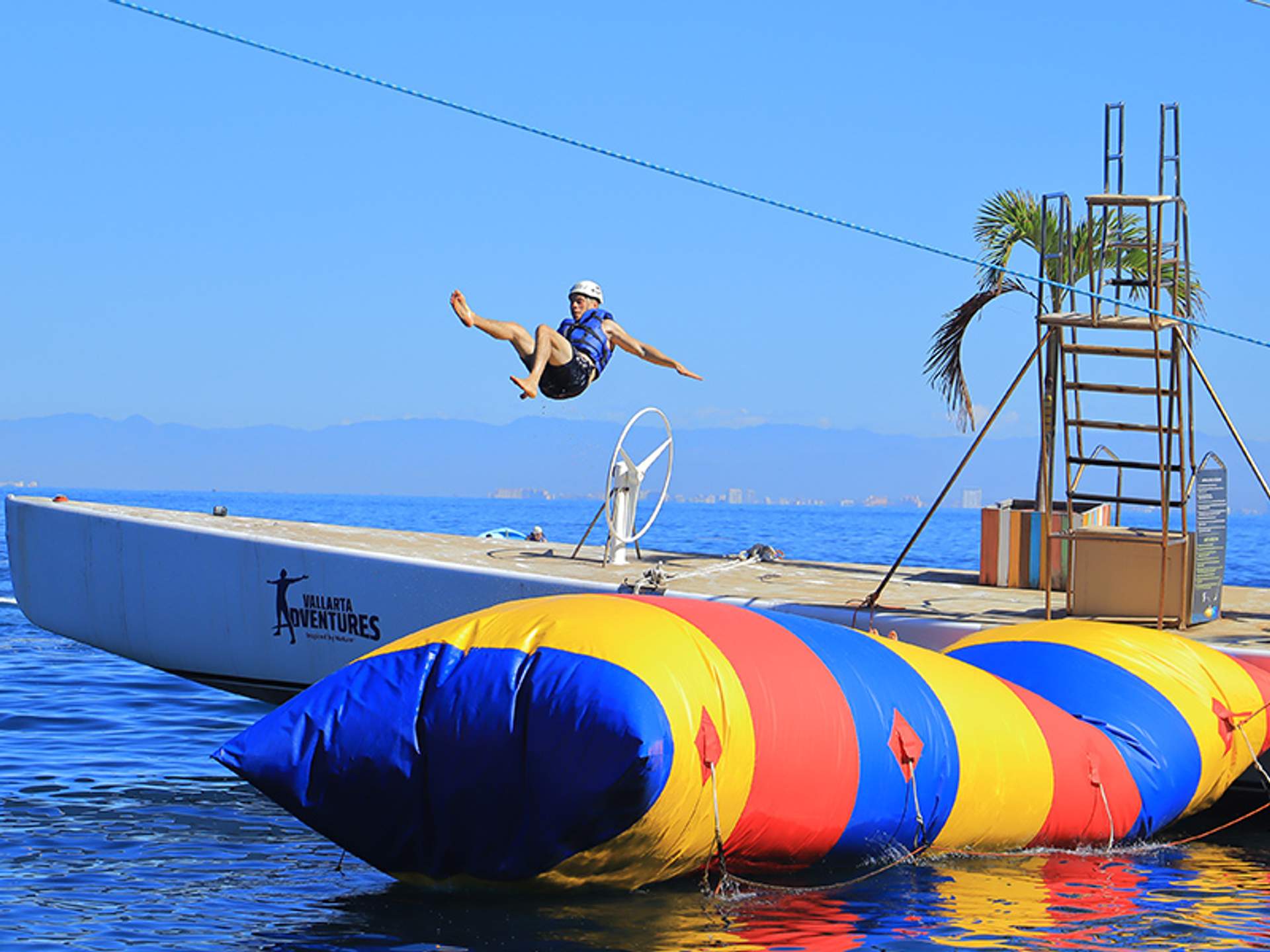 A person wearing a helmet and life jacket is mid-air above a colorful inflatable blob at an ocean water park, with mountains in the background.