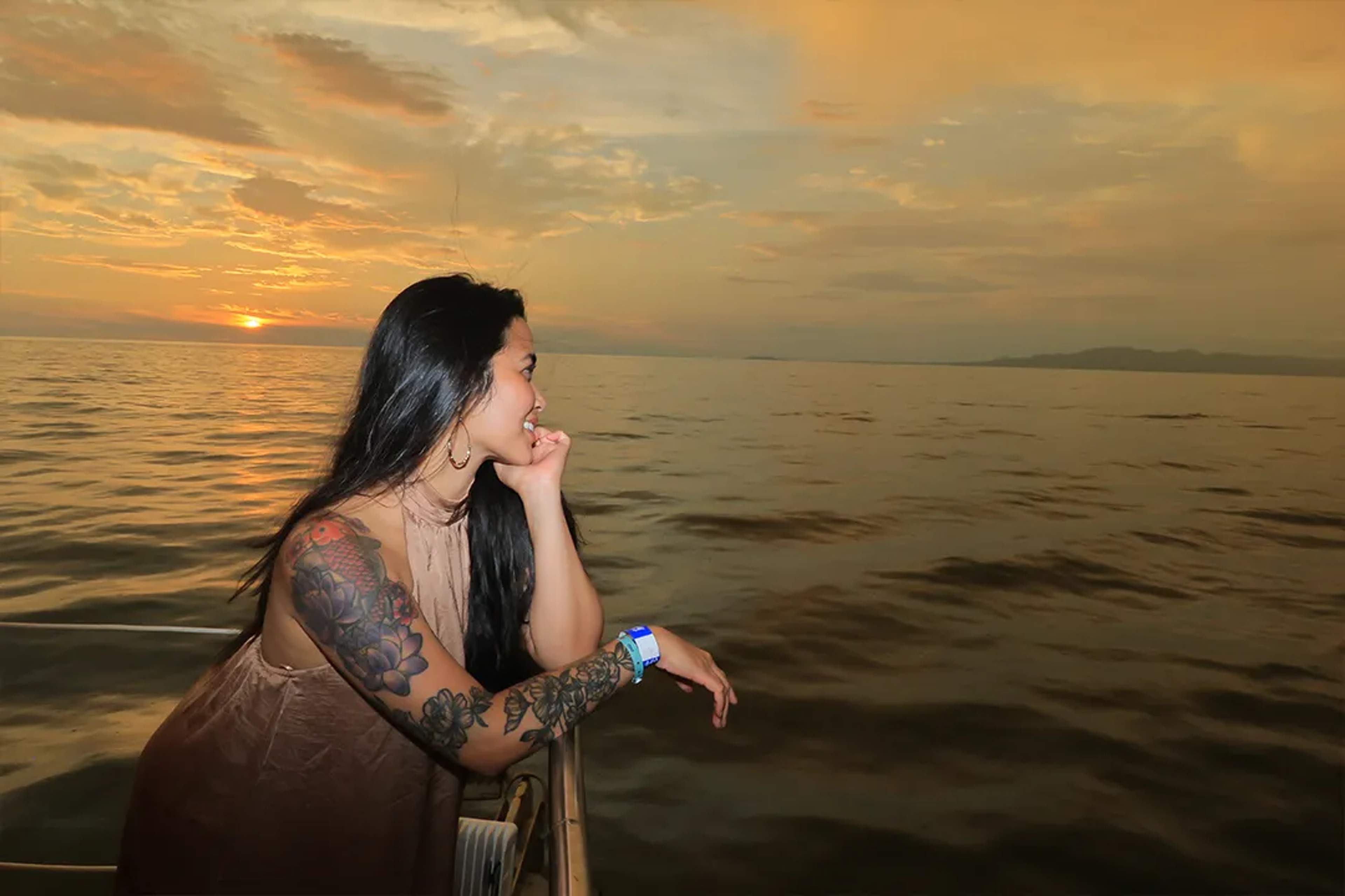 Woman with tattoos enjoys a peaceful sunset at sea, leaning on the boat railing.