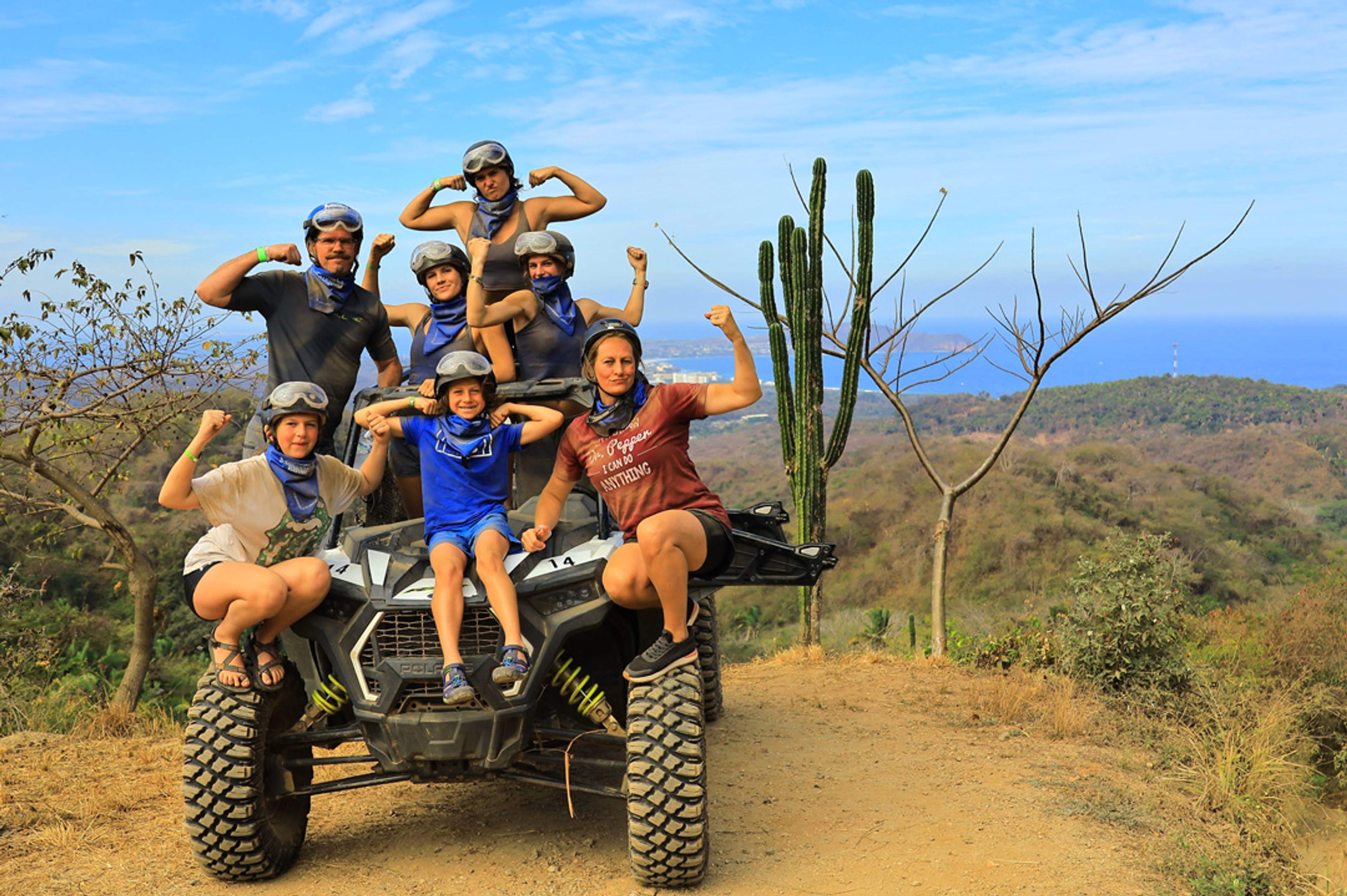 A group of people, including adults and children, pose flexing their arms on an all-terrain vehicle with a scenic view of hills and the ocean in the background. They are wearing helmets and protective gear.