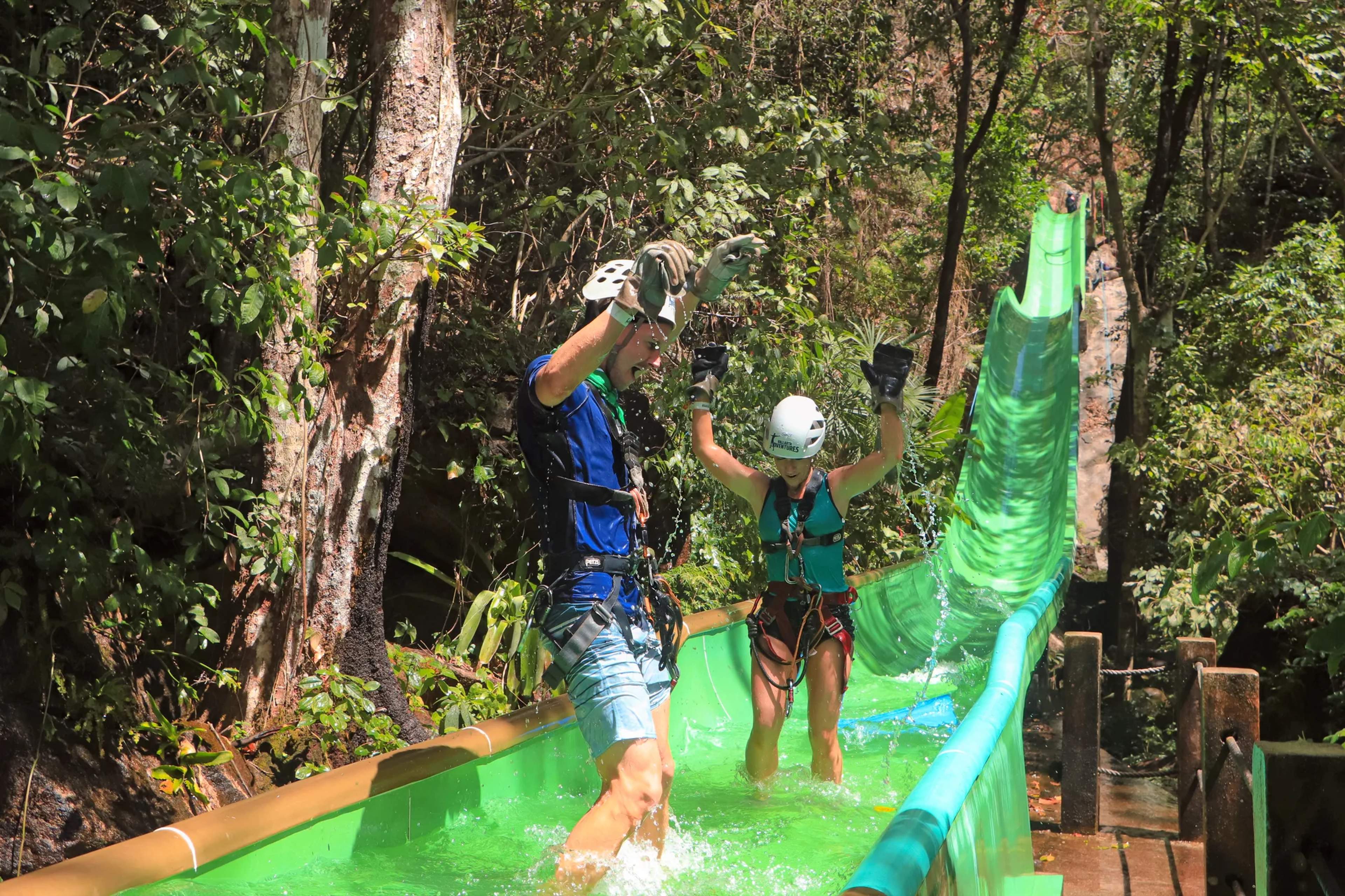 Adventurers zip line through a lush green jungle canopy on a bright suspended course above tall tropical trees.