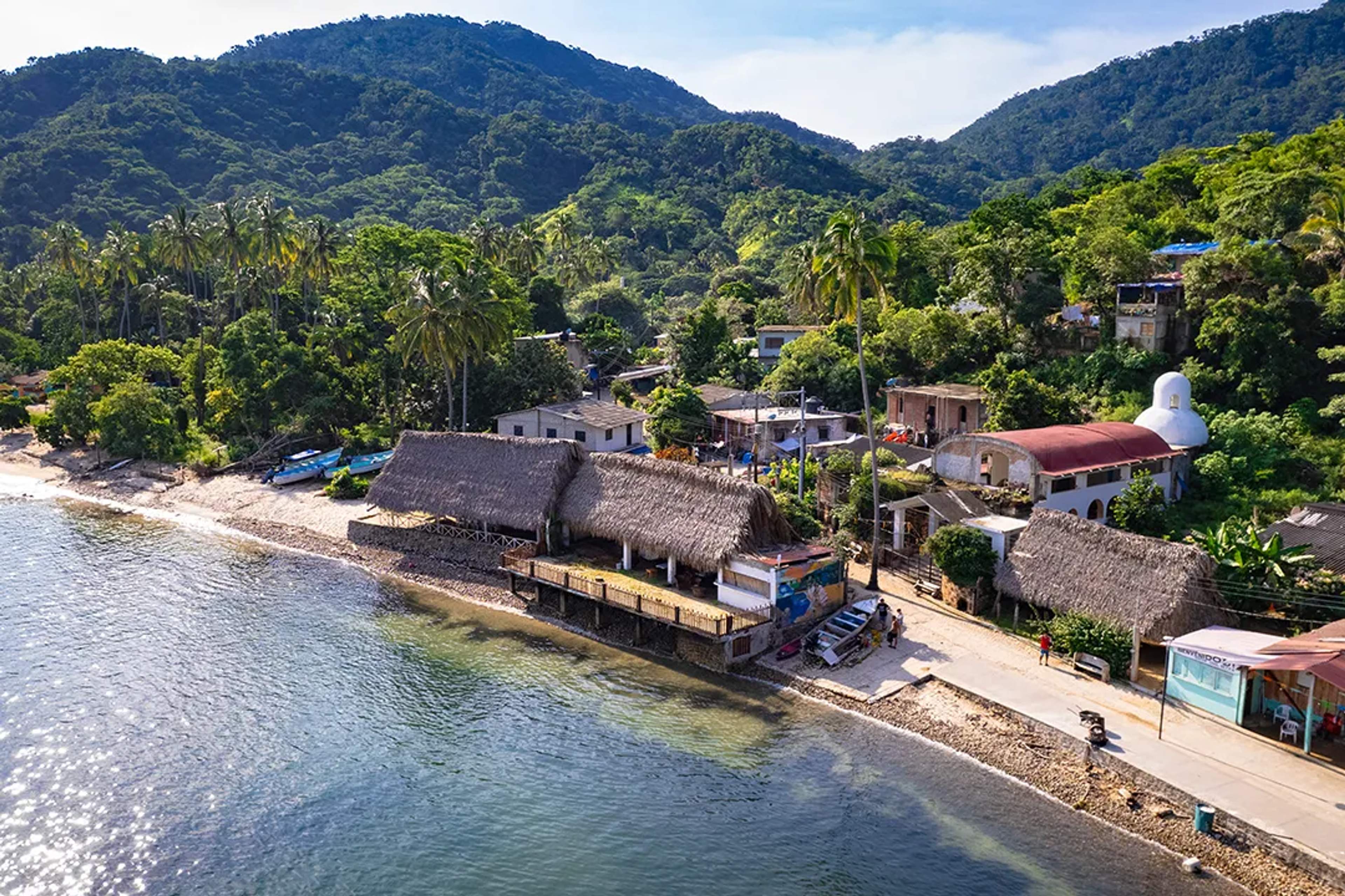 Vista aérea de un pueblo costero con techos de palapa, selva verde y mar tranquilo.