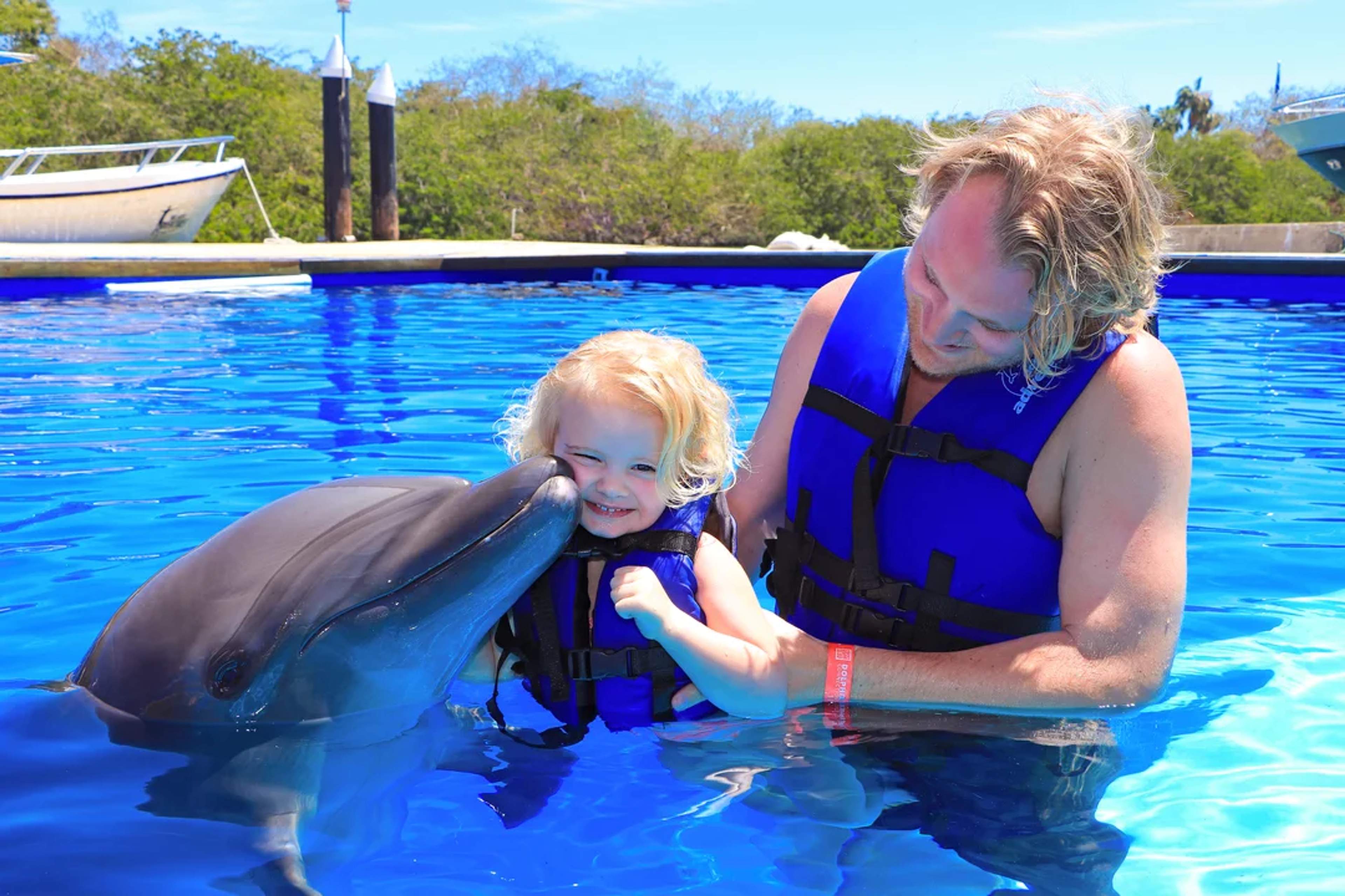 A dolphin kisses a smiling child in the water, creating a magical family moment.