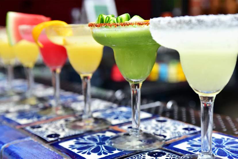Colorful margaritas in different flavors lined up on a tiled bar, garnished with fruit and chili.