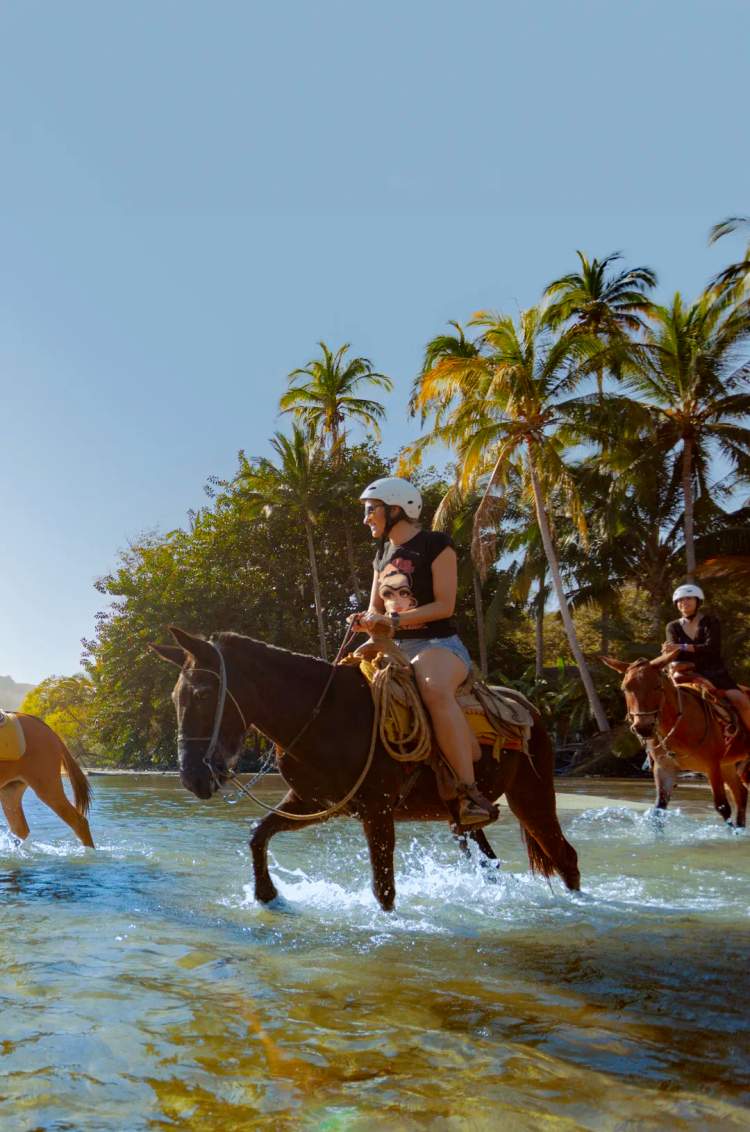 People horseback riding through the Yelapa river with lush greenery and mountains in the background.