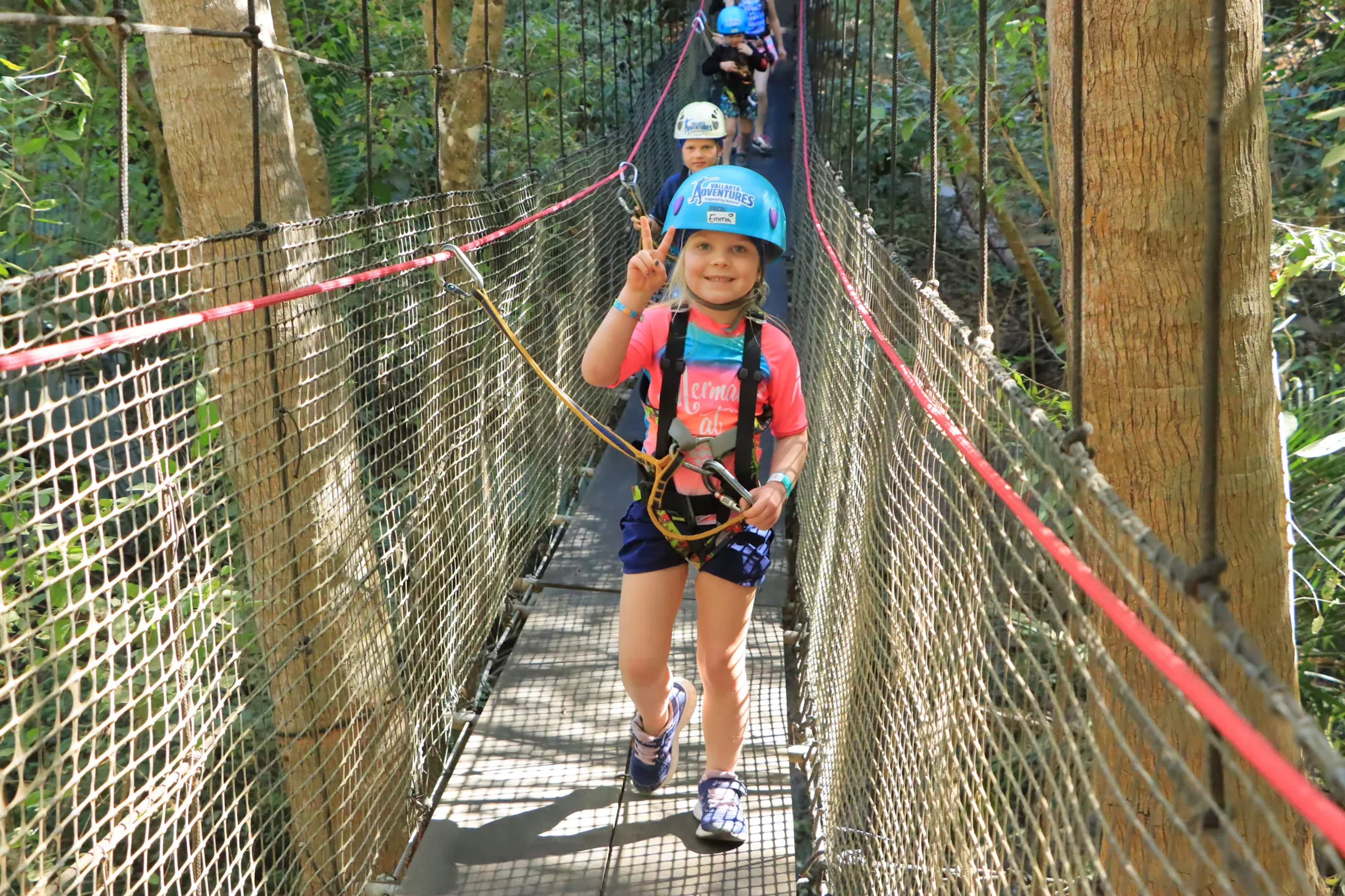 Niño pequeño con equipo de seguridad cruzando un puente colgante rodeado de árboles altos en la selva.