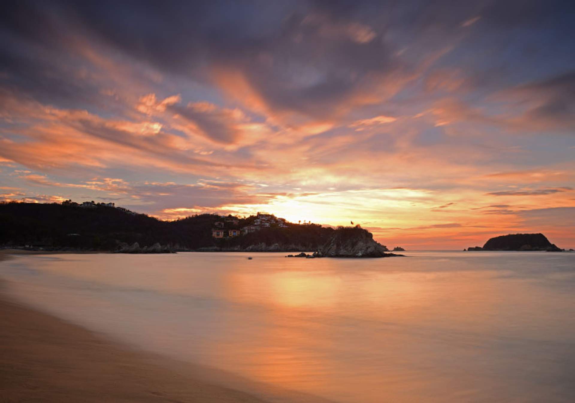 A stunning sunset over a tranquil beach, with the sky painted in vibrant shades of orange, pink, and purple. Silhouetted hills and rocky outcrops frame the serene ocean waters.