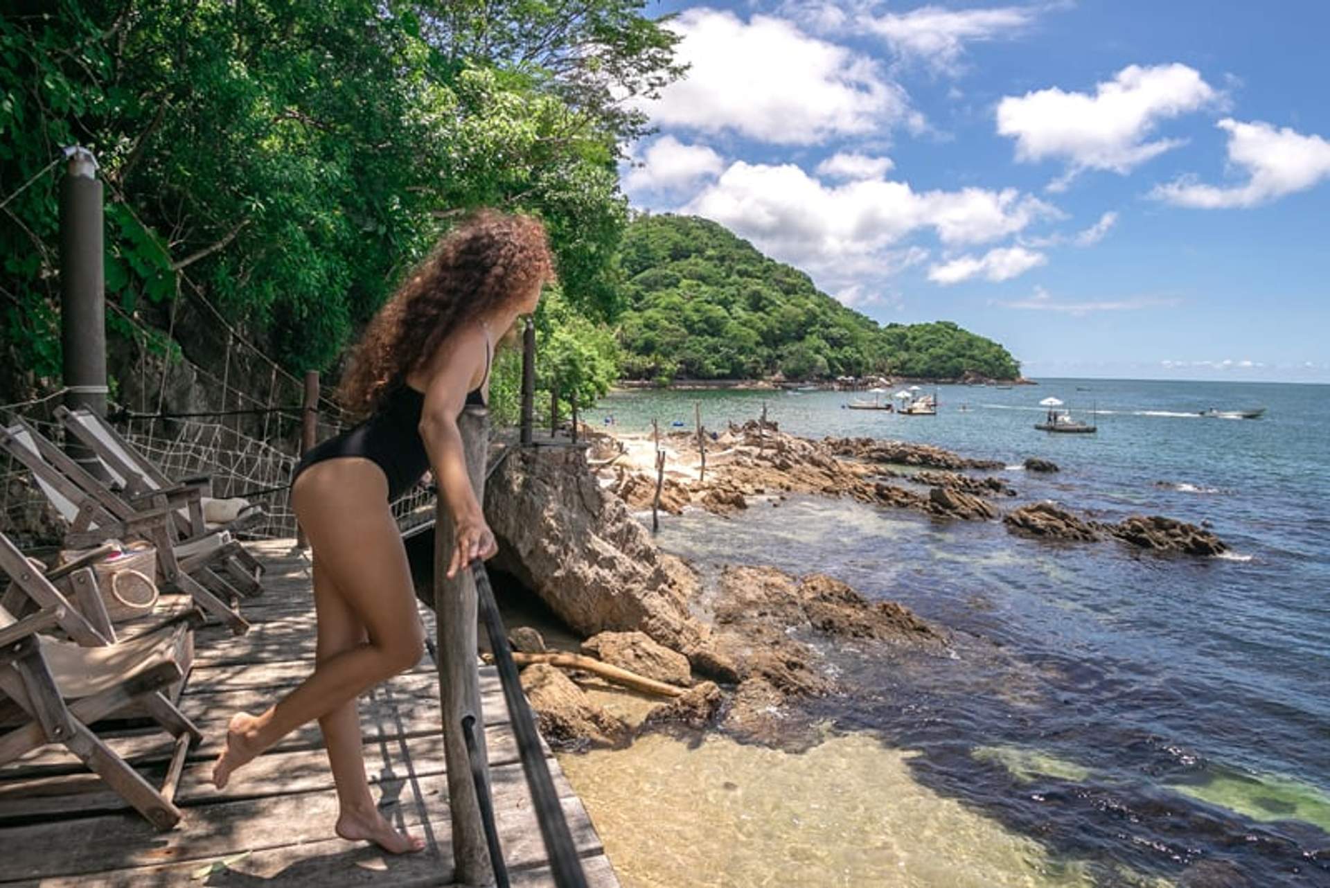 A woman in a black swimsuit standing on a wooden deck overlooking a rocky shoreline and the ocean. She is surrounded by lush greenery, and several boats are visible in the water under a bright blue sky.