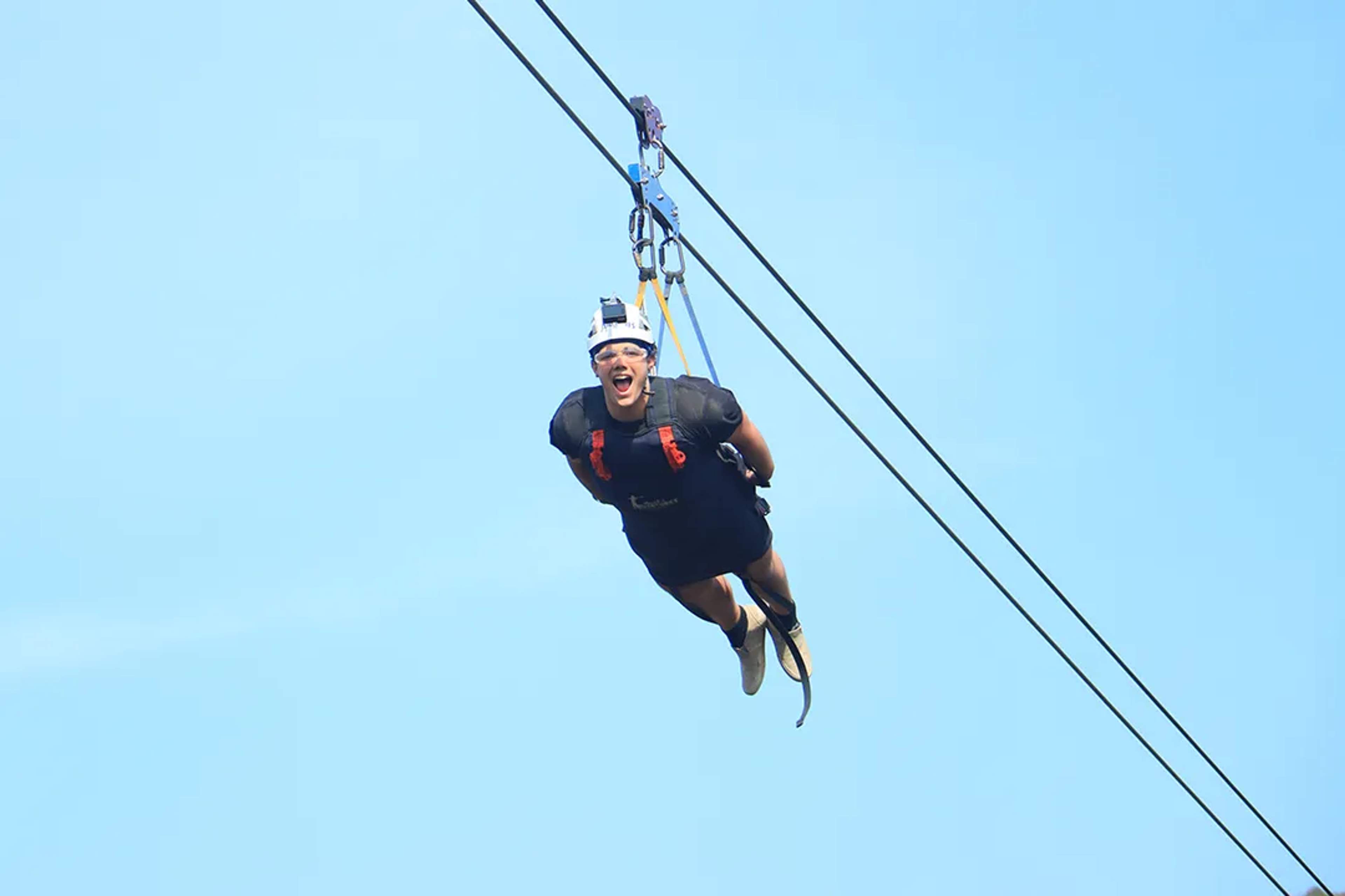 Adventurer soars Superman-style on a high-speed zipline, flying freely against the clear blue sky.