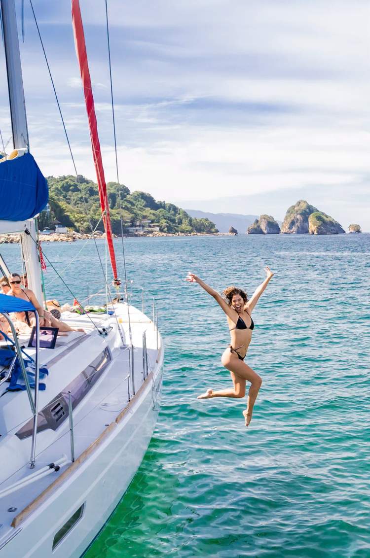 Woman jumping off a sailboat into the sea on the south shore of Puerto Vallarta with Los Arcos in the background.