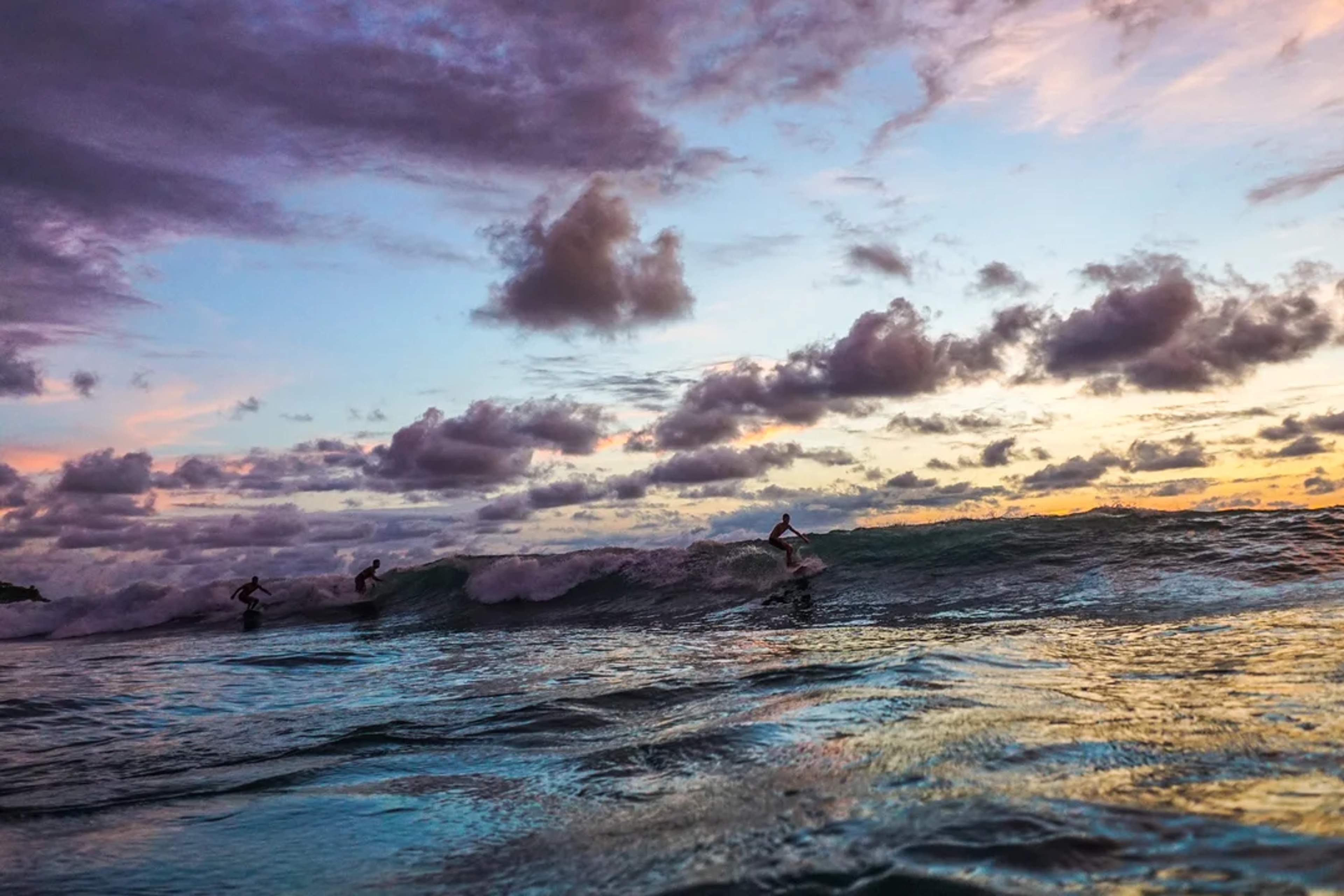 Surfistas montando olas al atardecer bajo un cielo colorido y lleno de nubes dramáticas.