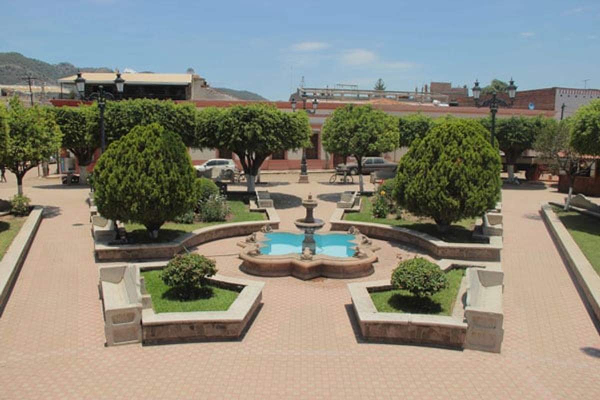 Central plaza in Mascota, Jalisco, with a fountain in the center, pruned trees, stone benches, and colonial buildings in the background.