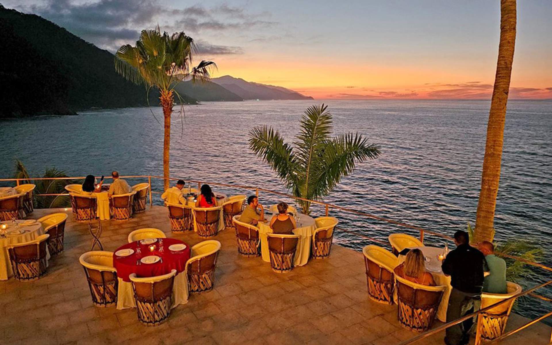 Diners enjoying a sunset meal on a terrace overlooking the ocean, with palm trees and mountains in the background.