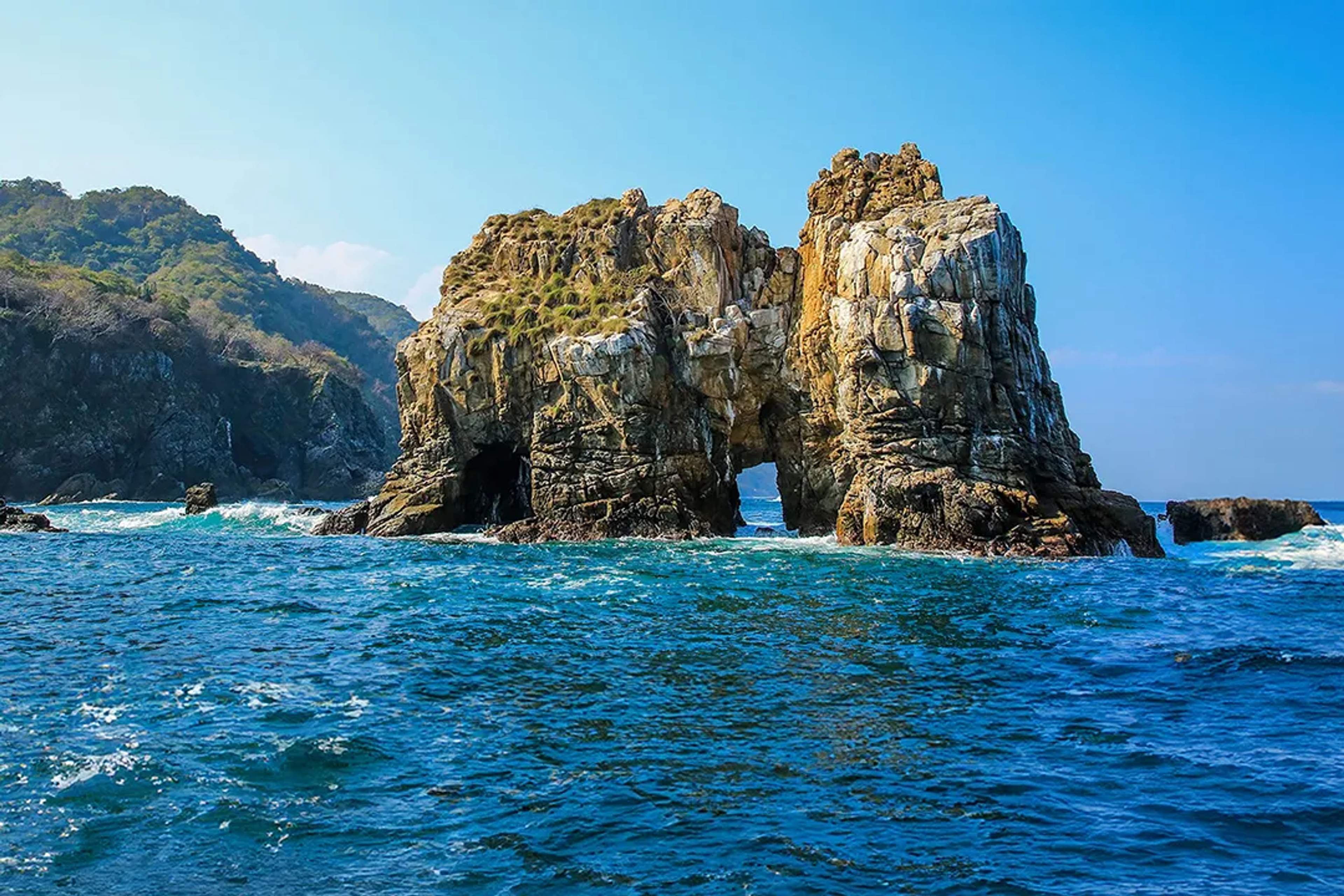 Rocky islet with natural arches rises from the Pacific, surrounded by deep blue waters and waves.
