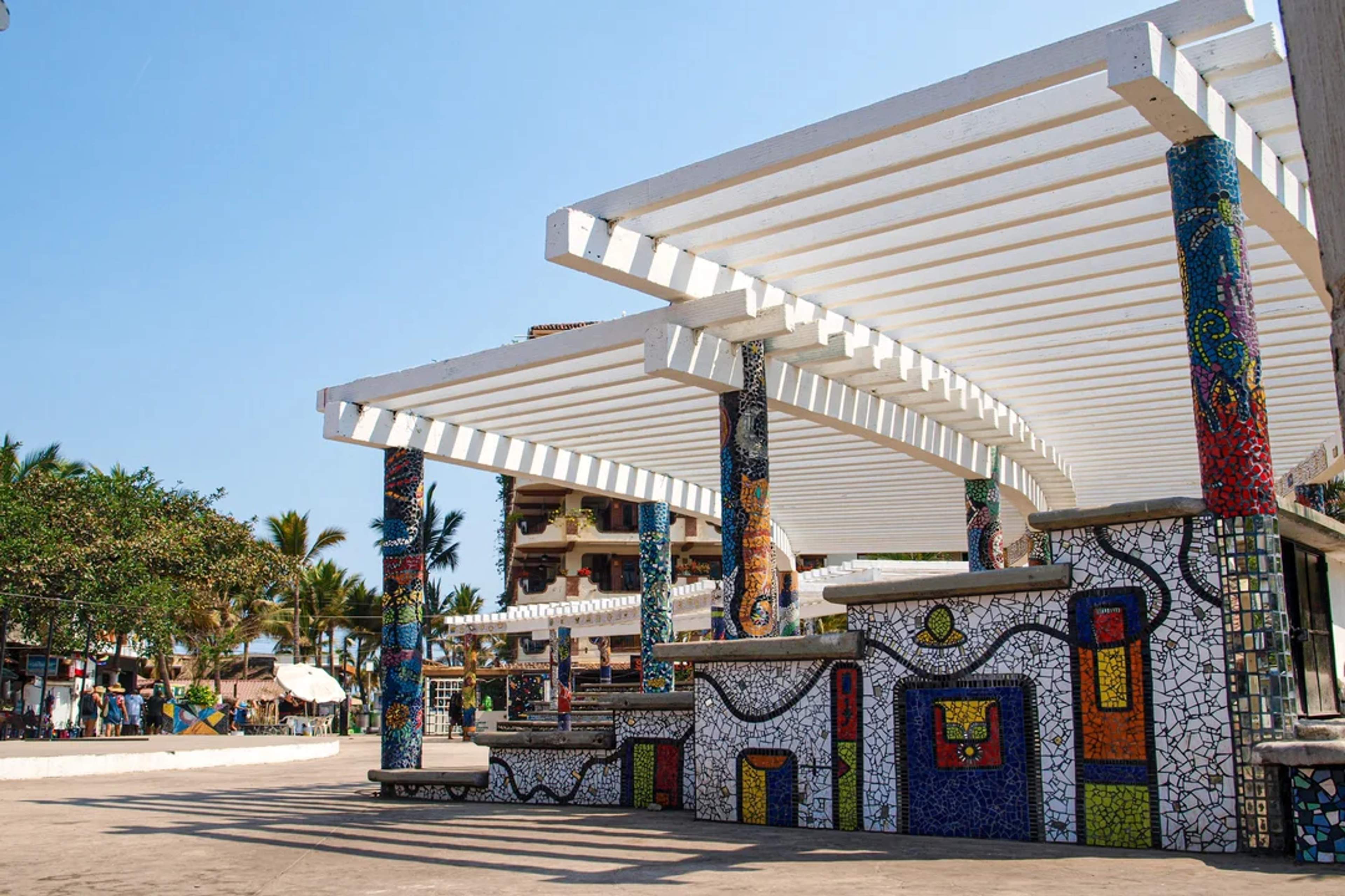 Colorful mosaic columns and benches decorate a shaded public space in a sunny coastal town.