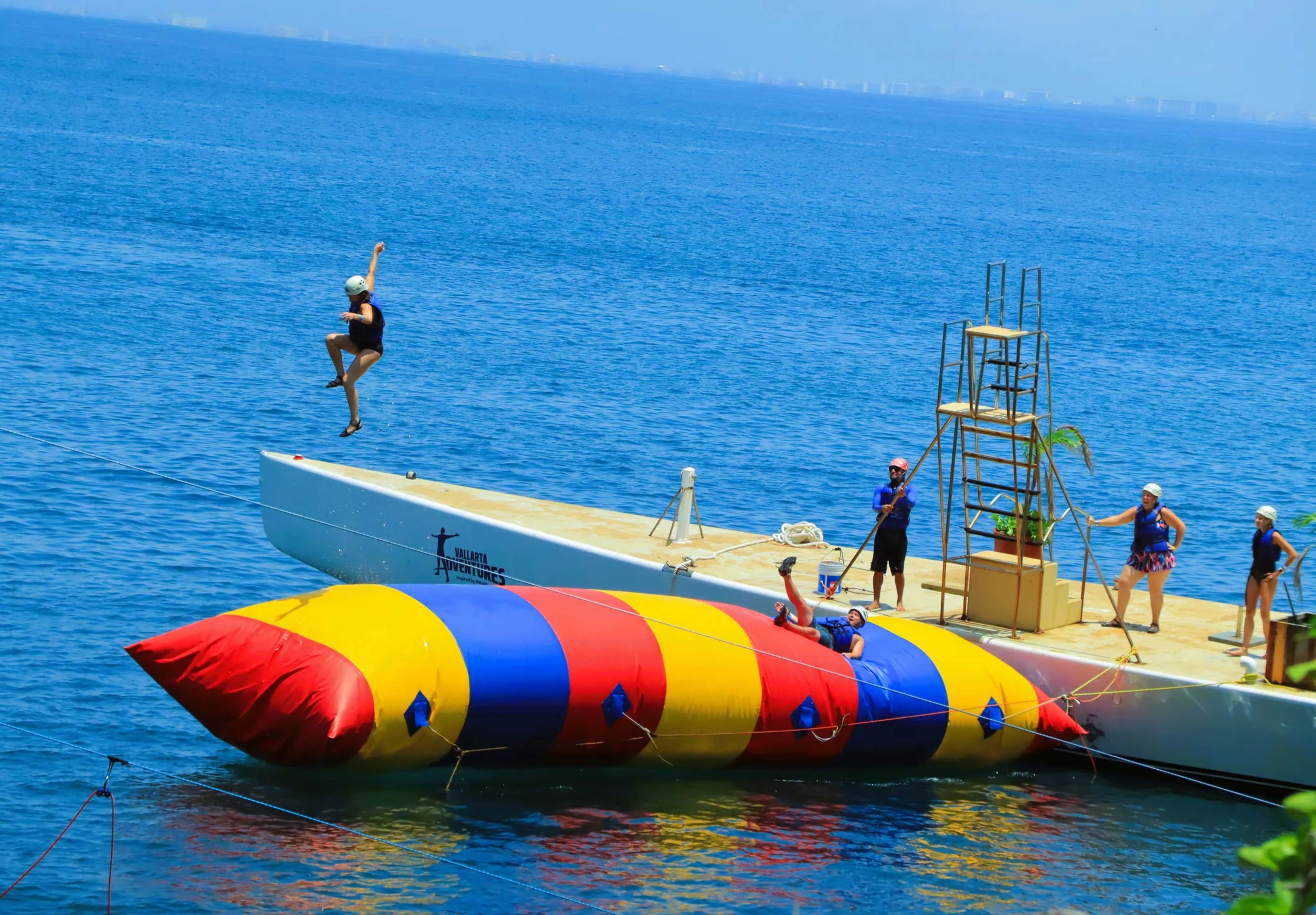 Tourist leaps off a white boat into crystal-clear blue ocean water near a colorful inflatable toy during an adventure tour.