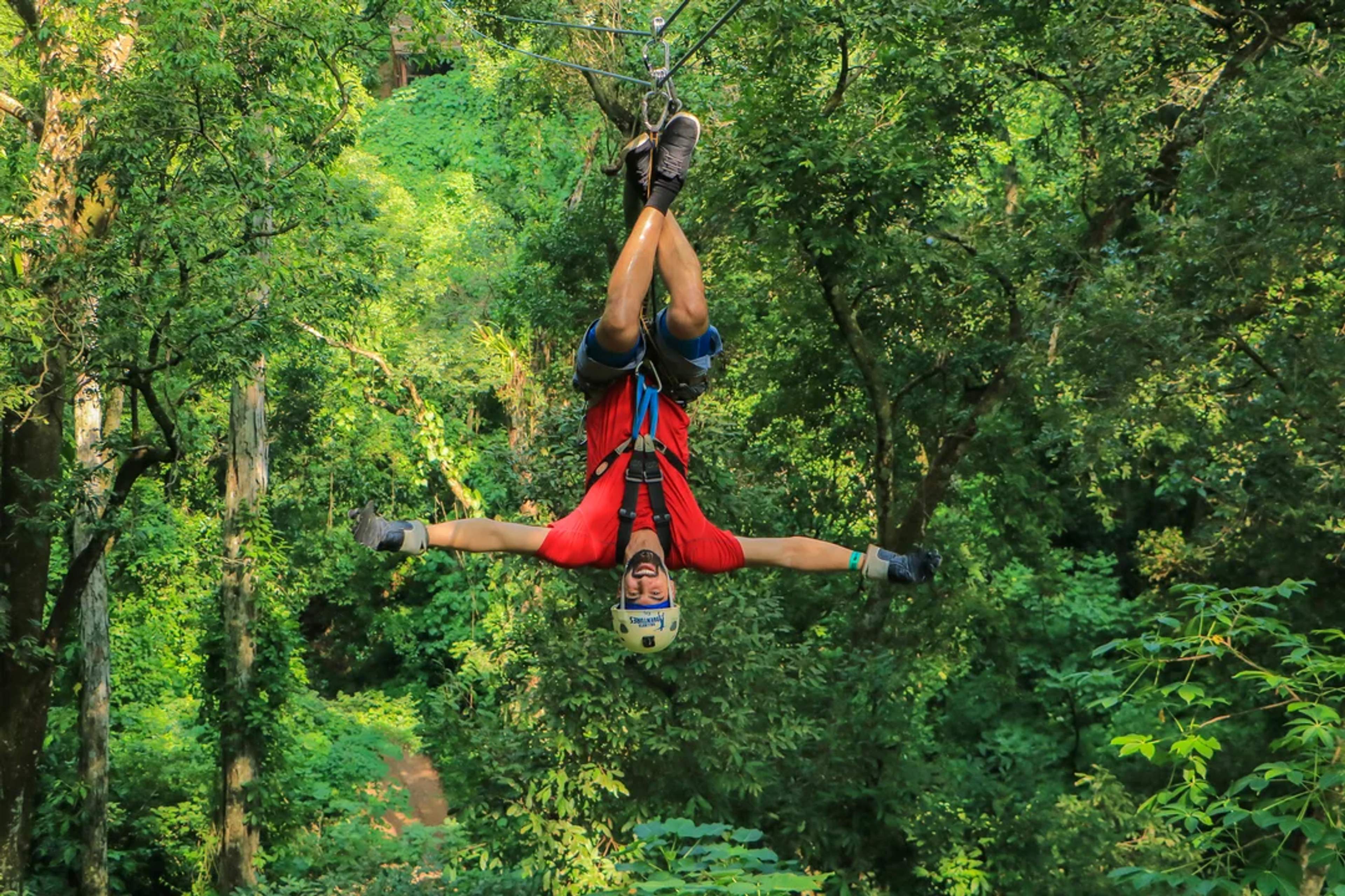 Adventurer ziplines upside down through lush jungle, arms wide open and full of excitement.