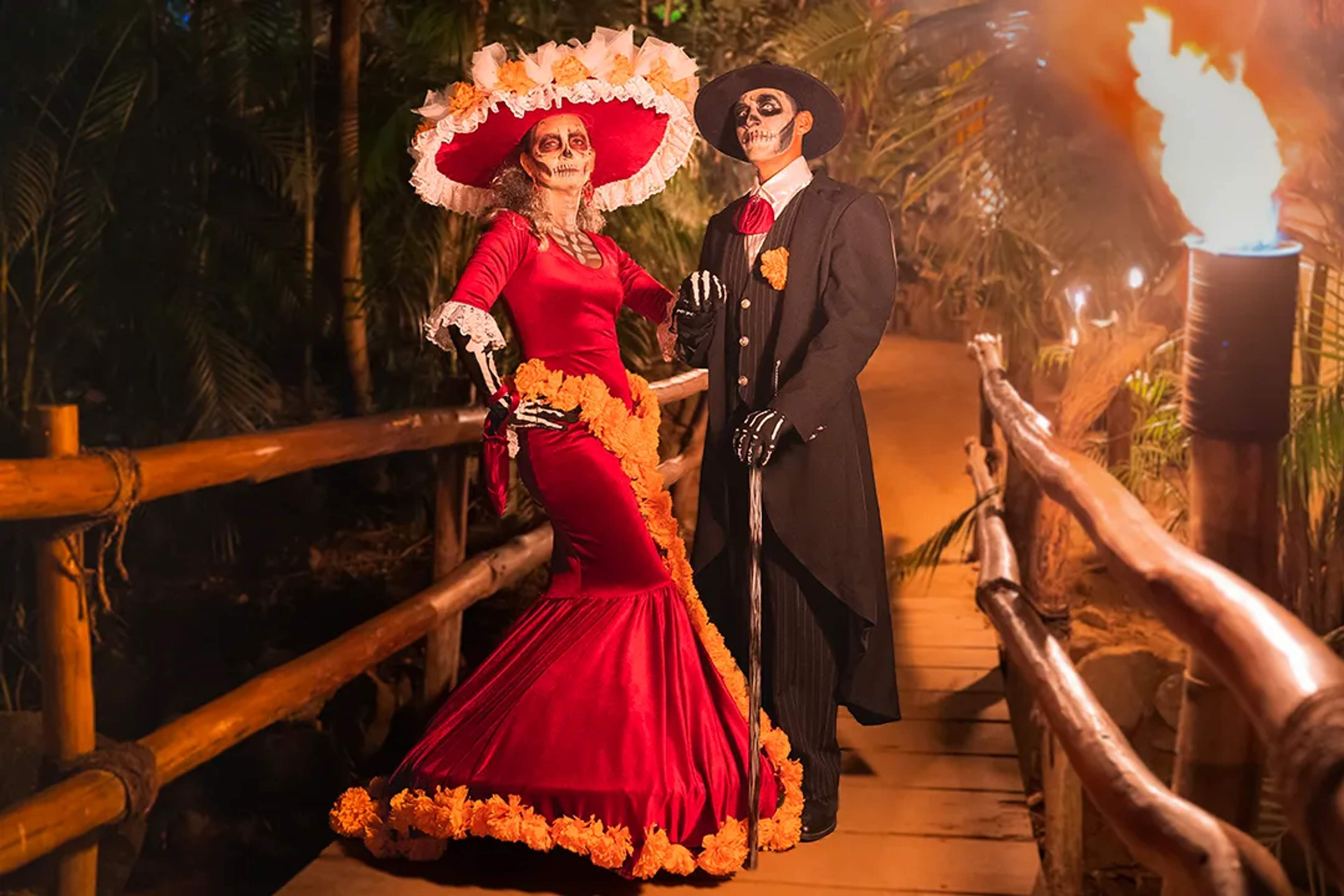 Elegante pareja de Día de Muertos con trajes de Catrina sobre un puente de madera iluminado.