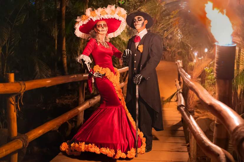 Elegant Day of the Dead couple in traditional Catrina attire on a candlelit wooden bridge.