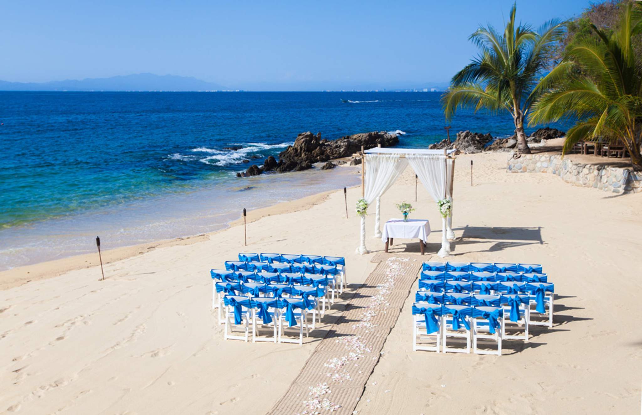 Configuración de boda en la playa con sillas blancas adornadas con cintas azules frente a un altar junto al océano en Puerto Vallarta, México.