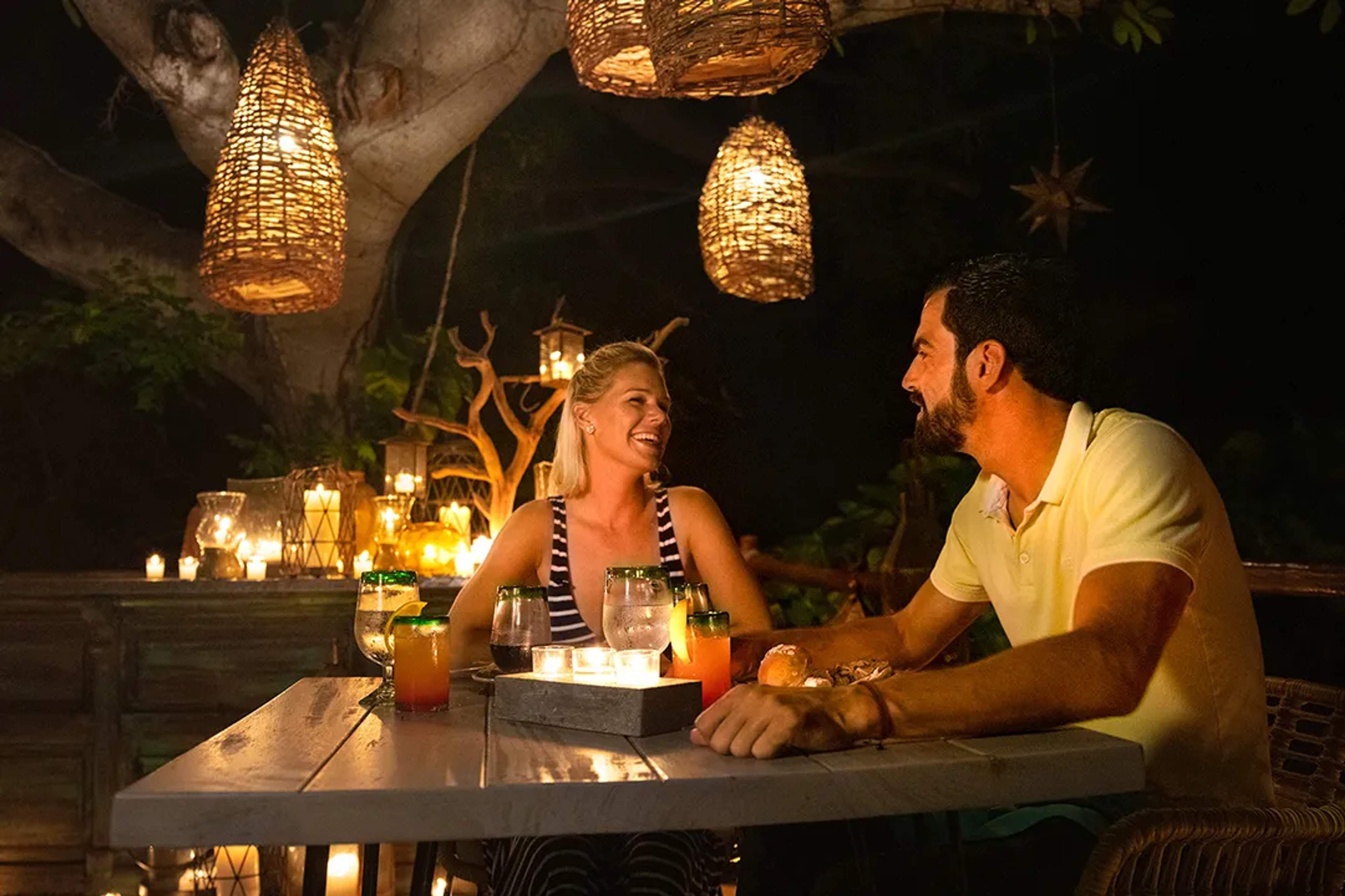 Pareja disfruta una cena romántica al aire libre iluminada por velas y linternas colgantes.