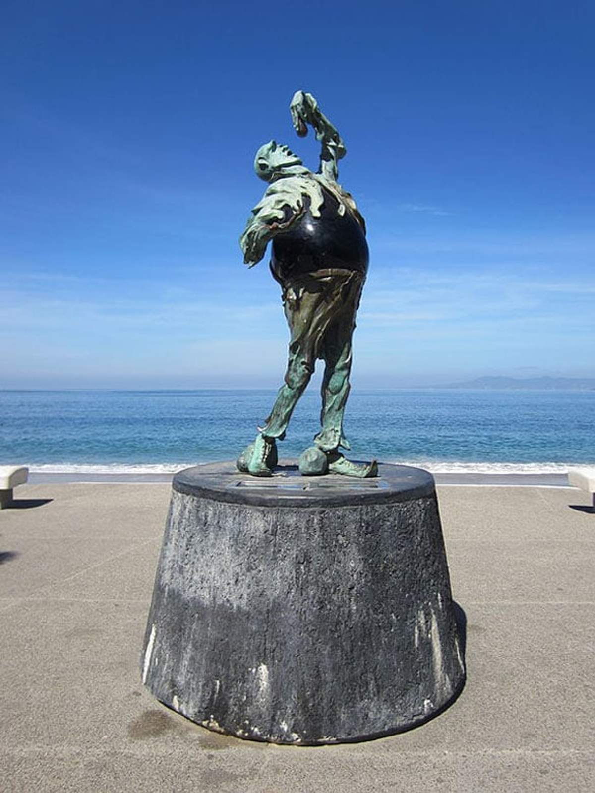 Escultura en el malecón de Puerto Vallarta, figura abstracta sobre pedestal con el océano y cielo despejado de fondo.