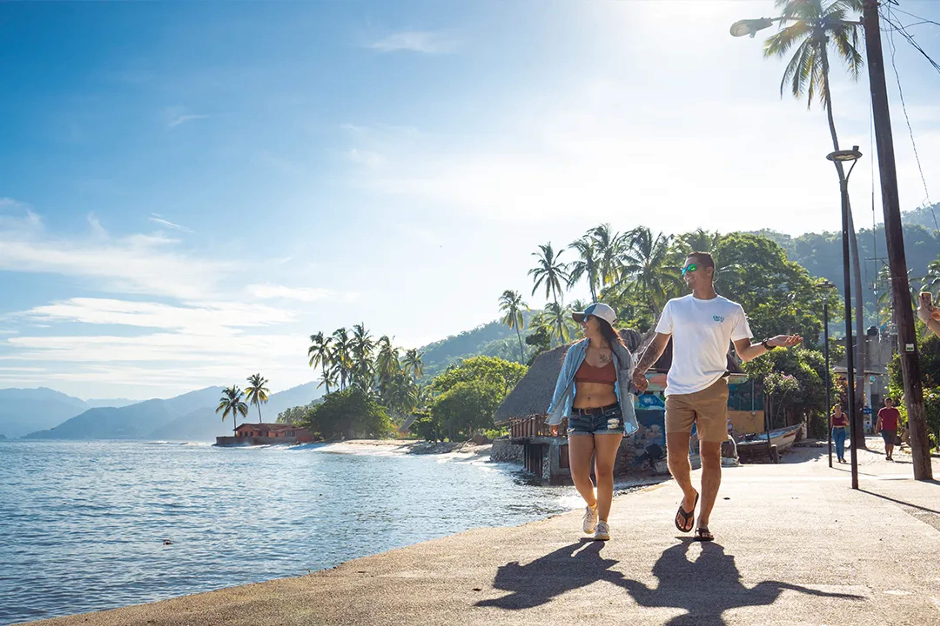 Pareja camina junto al mar en un día soleado, rodeada de palmeras y montañas verdes.