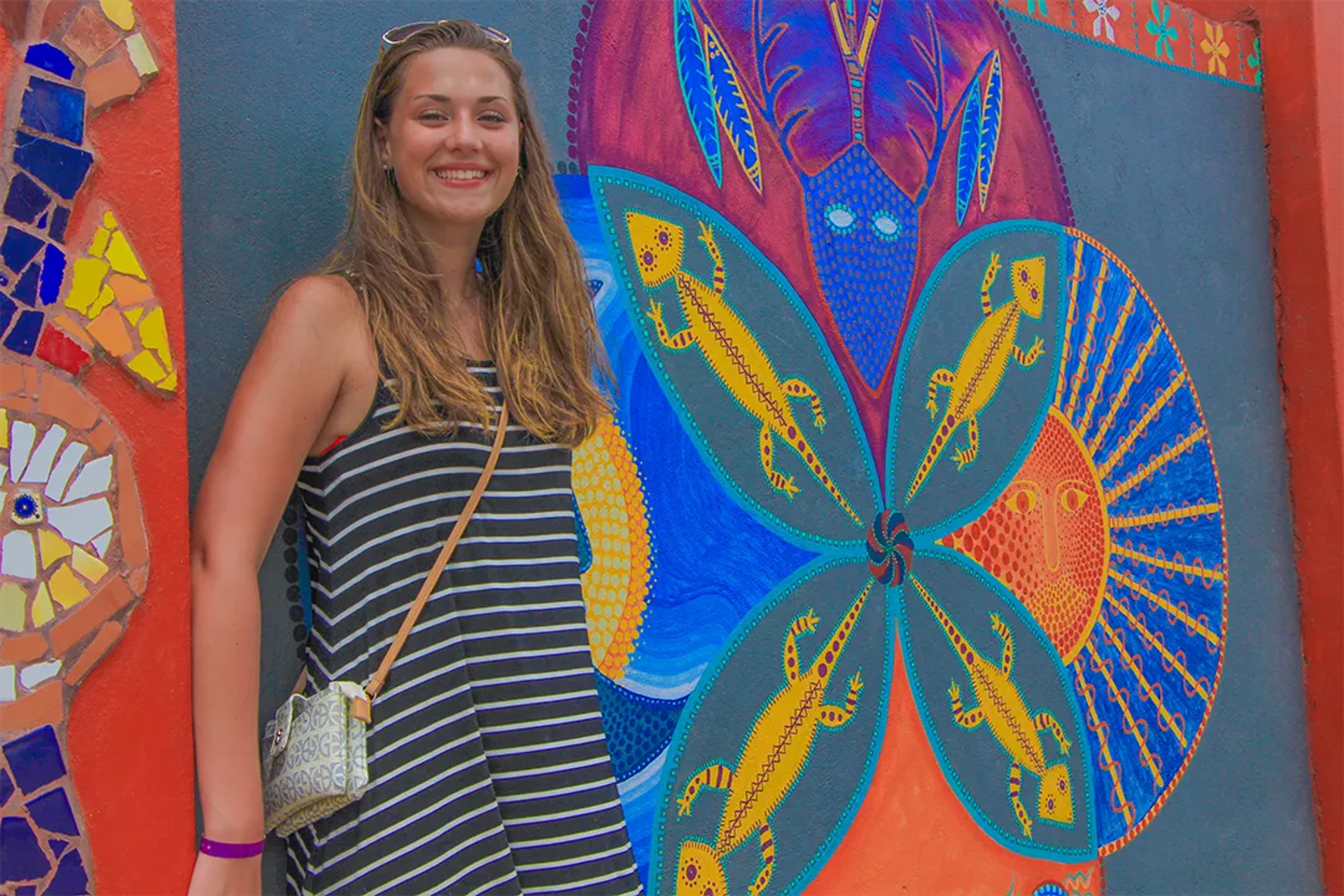 Smiling woman poses by colorful Mexican mural featuring lizards, eyes, and vibrant patterns.