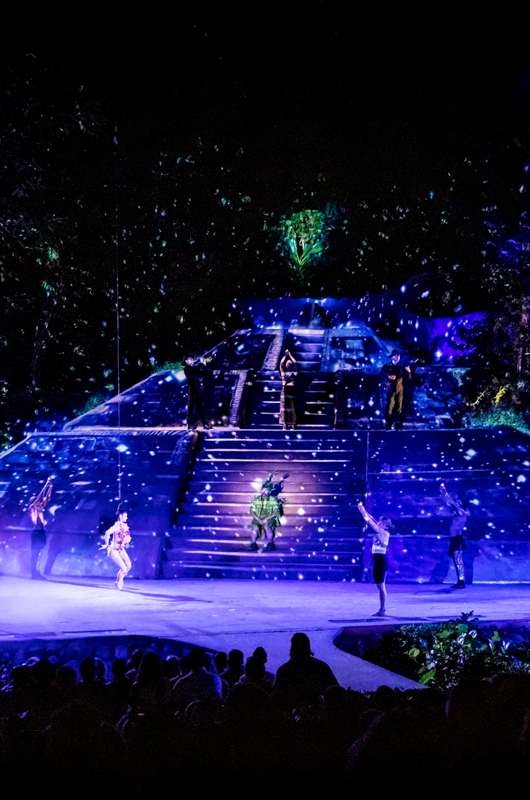 A vibrant night performance on an open stage surrounded by tropical jungle, with dancers illuminated by colorful lights.