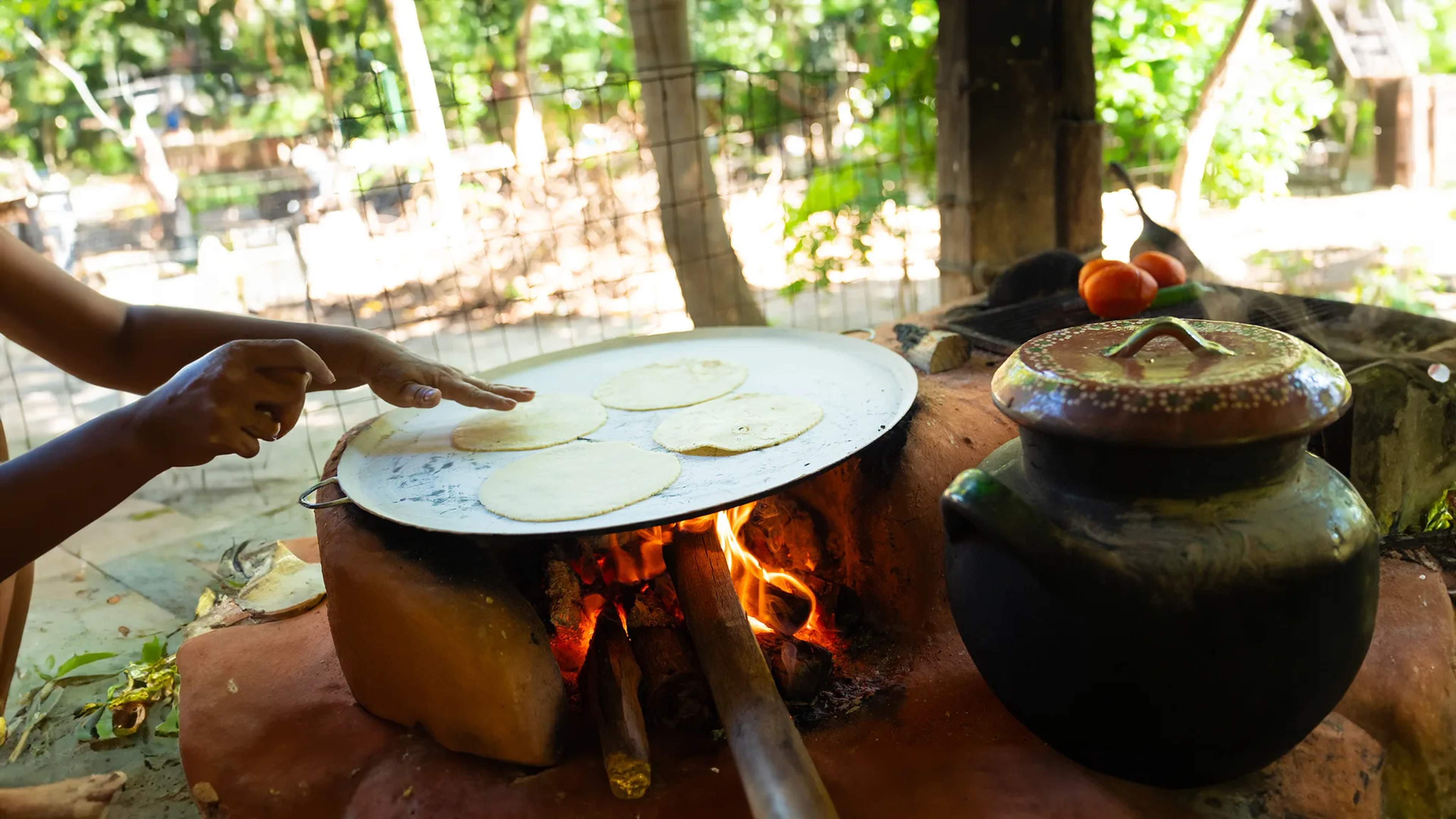 Una cocinera prepara tortillas a mano sobre fuego de leña en una cocina rústica al aire libre en un pueblo tropical