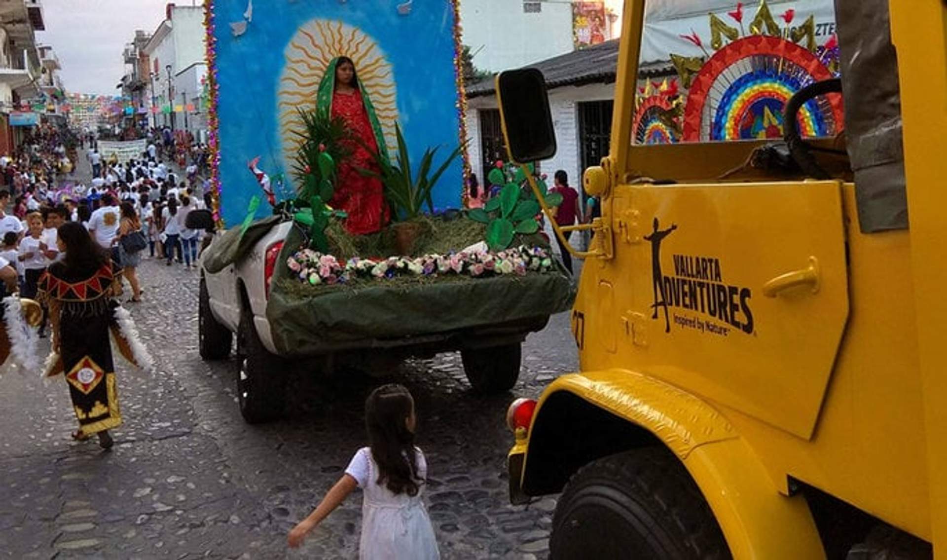 Carro alegórico de la Virgen de Guadalupe en Puerto Vallarta, con una niña caminando hacia él y decoraciones coloridas alrededor.