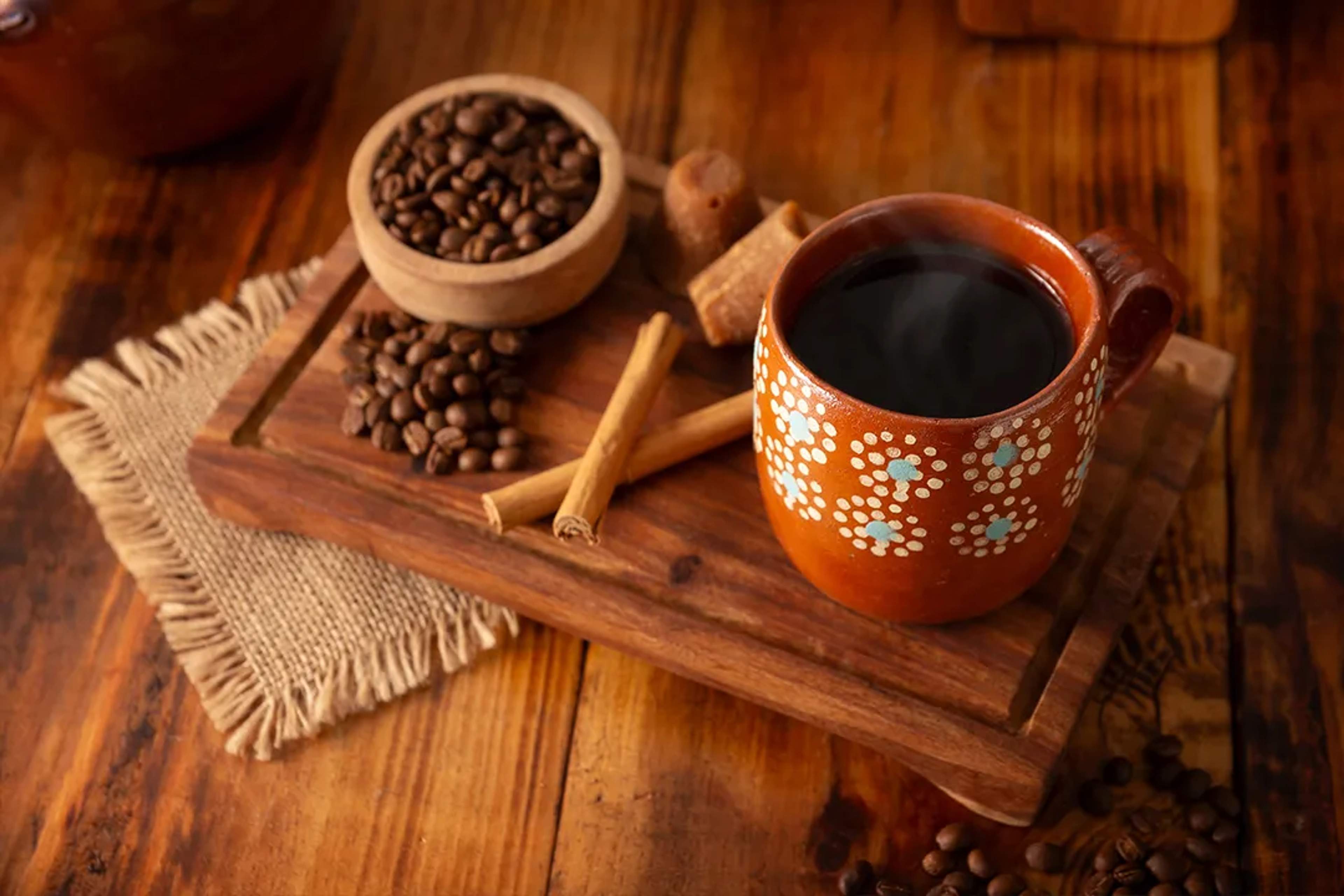 Jarro de barro con café de olla, canela, piloncillo y granos de café sobre mesa rústica.